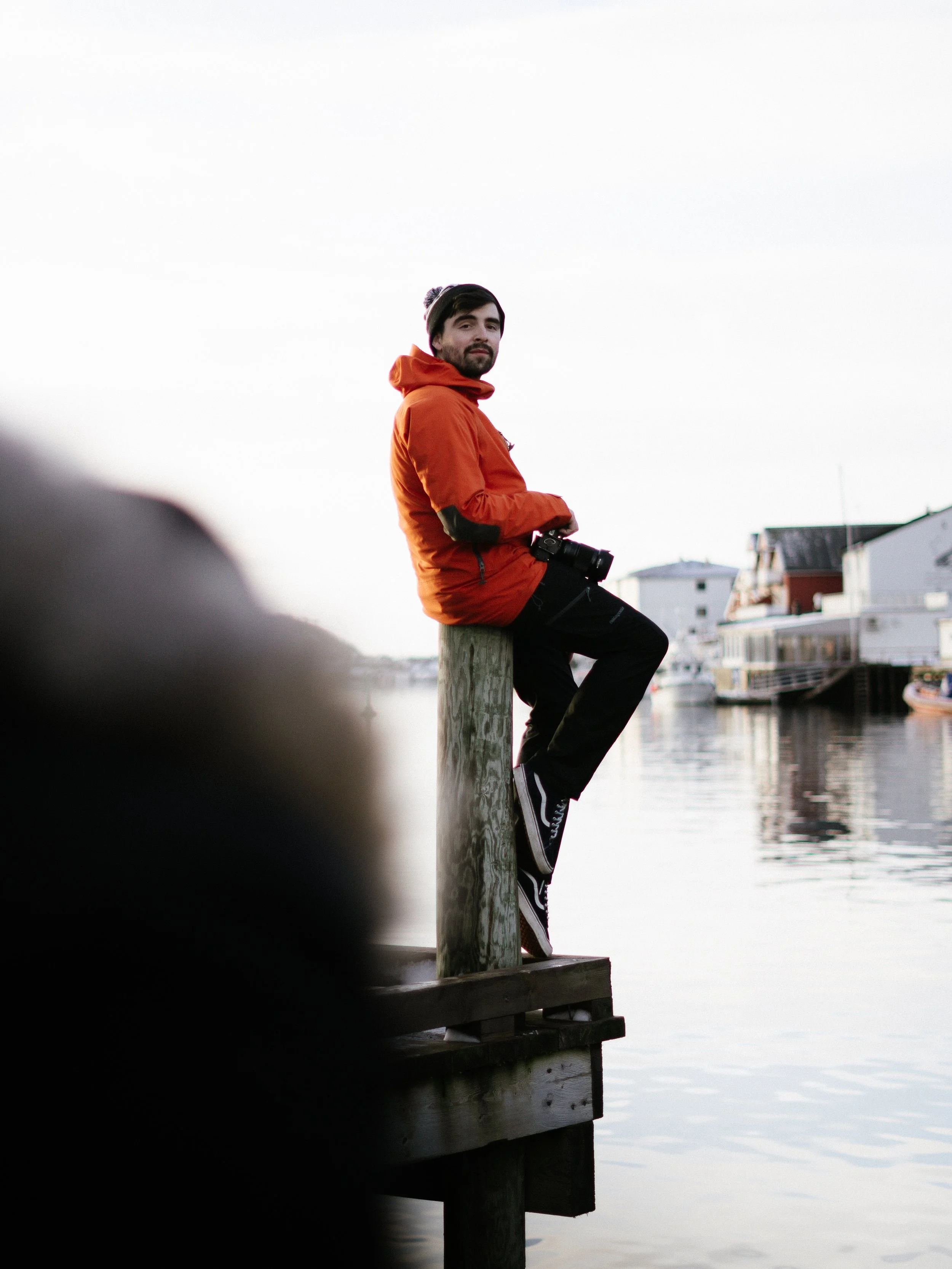 A man with dark hair, beard, wearing an orange jacket, black pants, and a beanie, sitting on a wooden dock with a camera hanging around his neck, near a calm body of water, with houses in the background during early morning or late evening.