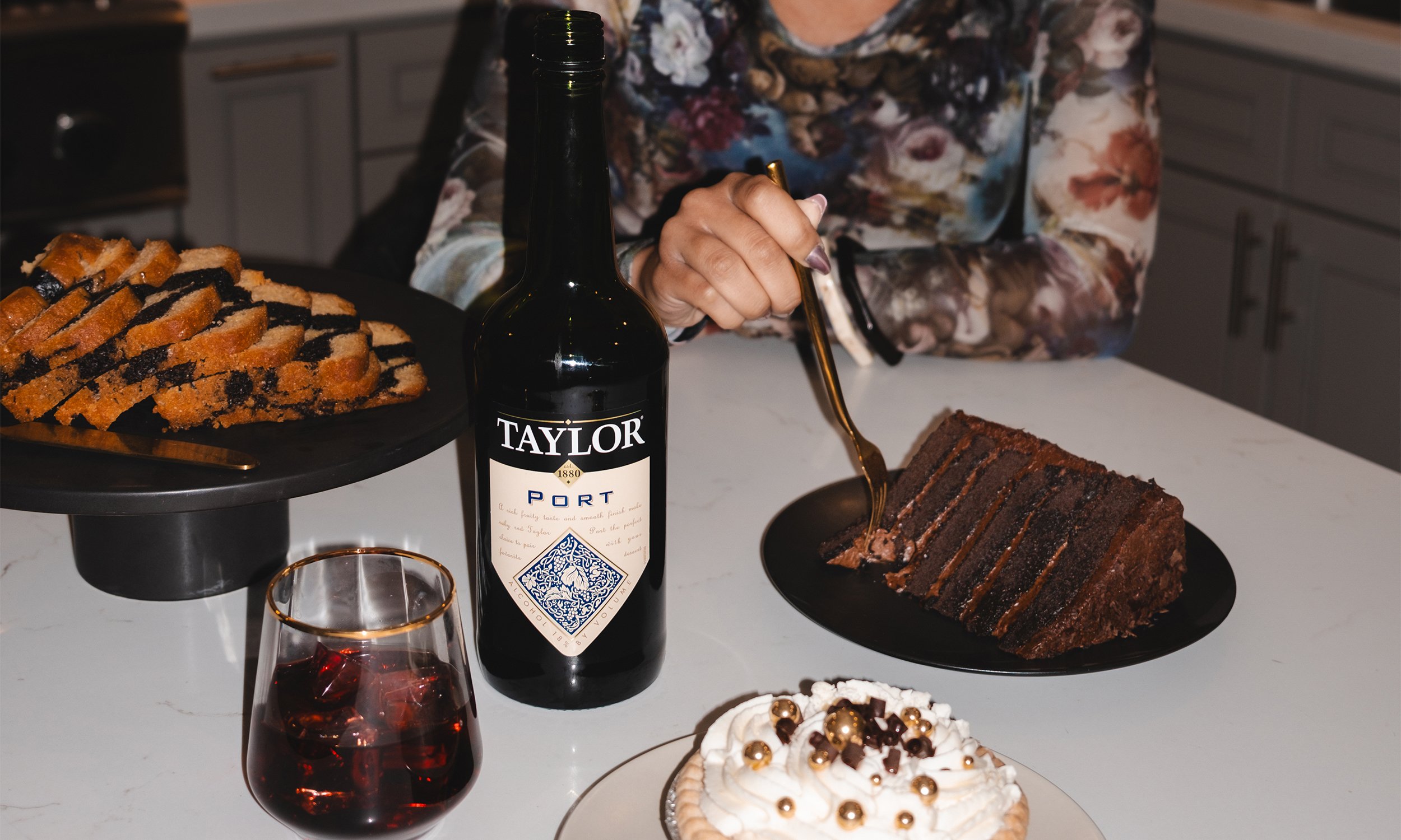 A dessert table with a chocolate cake, a topped cream pie with gold decorations, and a plate of assorted cookies. A person is pouring a drink, and there's a bottle of Taylor Port wine and a glass of red wine.