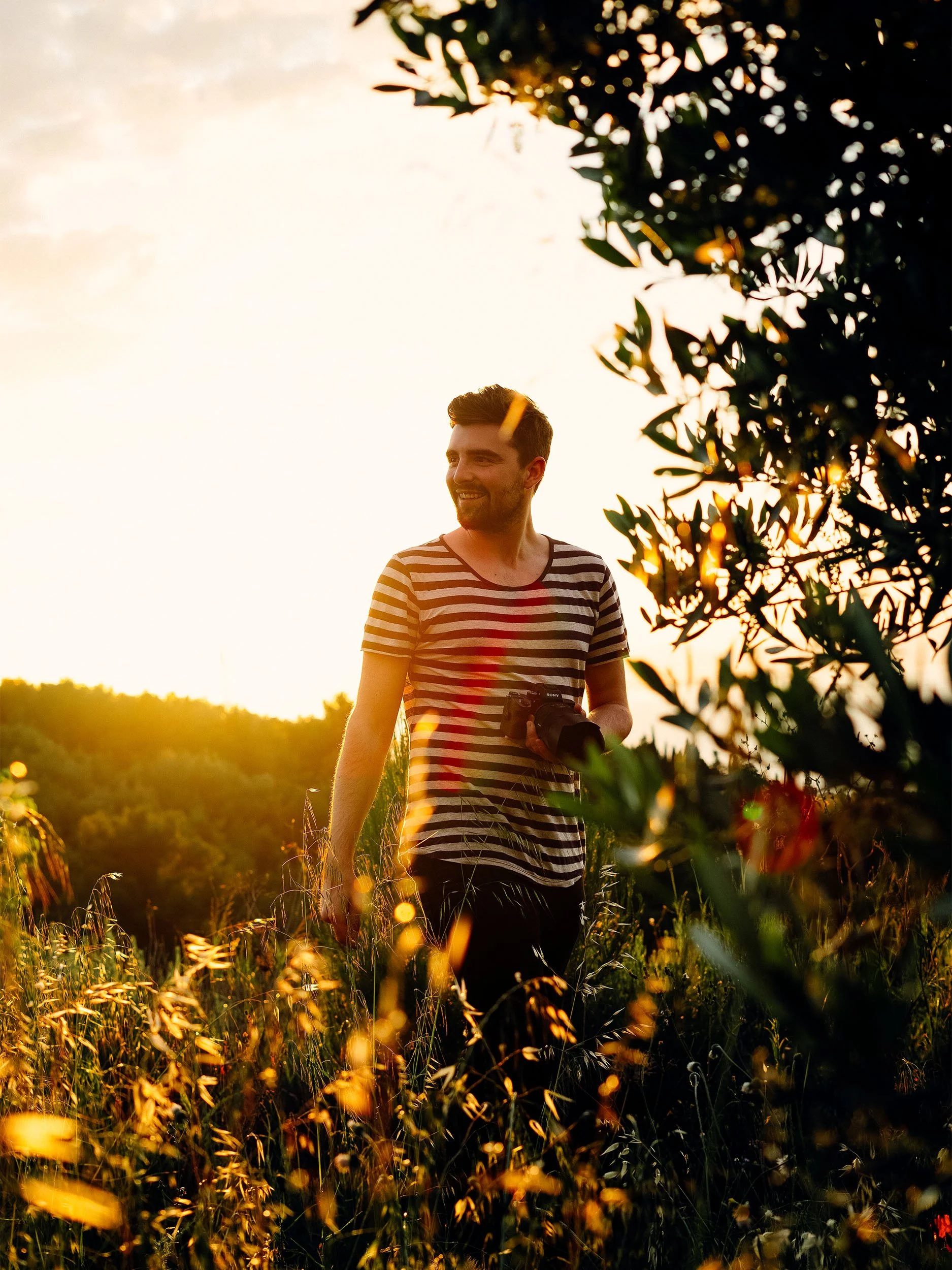 A man with a beard in a striped shirt holding a camera walking through a field at sunset, with the sunlight filtering through the leaves of trees.