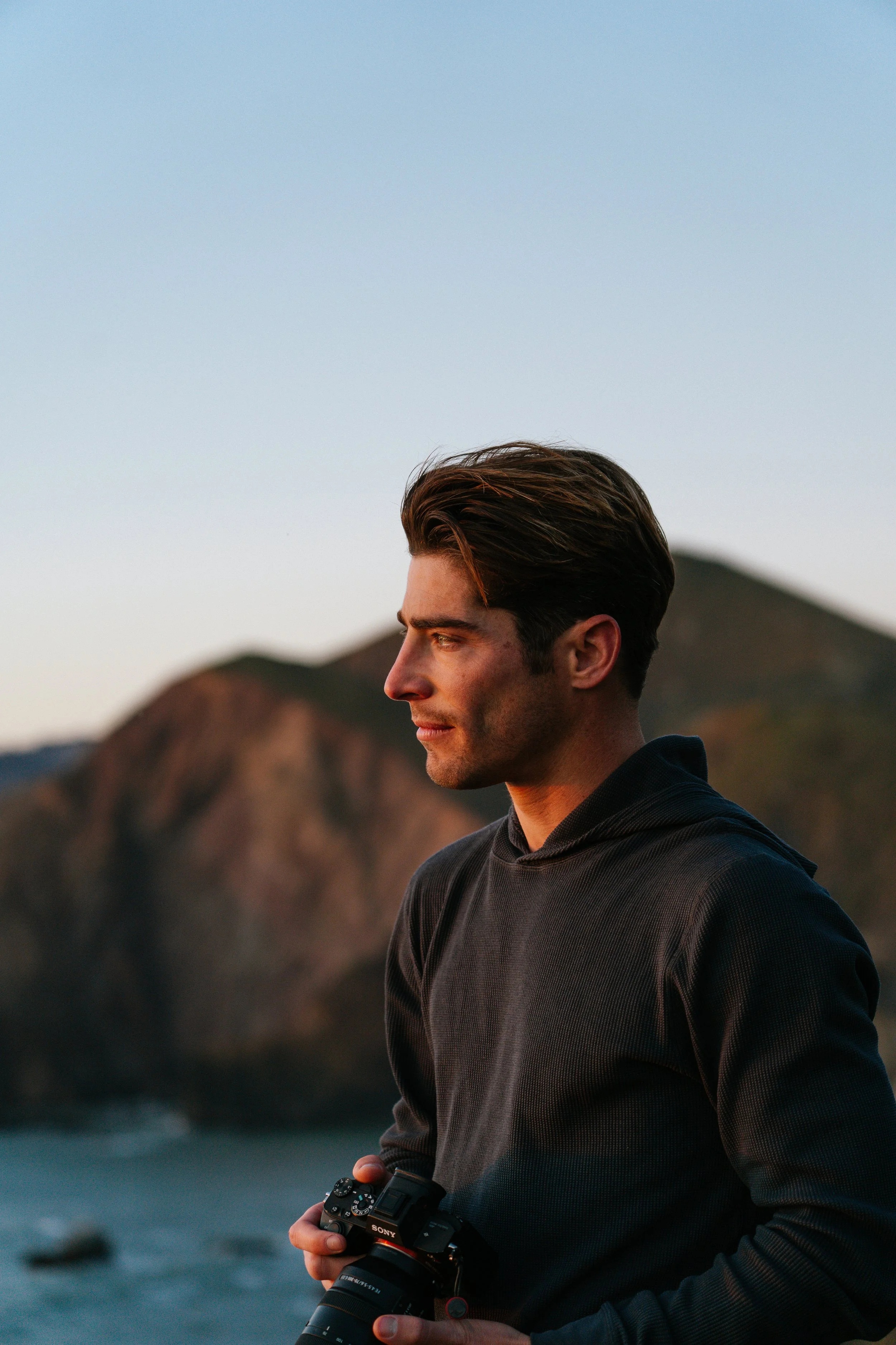 Young man with brown hair holding a camera while standing outdoors during sunset, with a mountain landscape in the background.