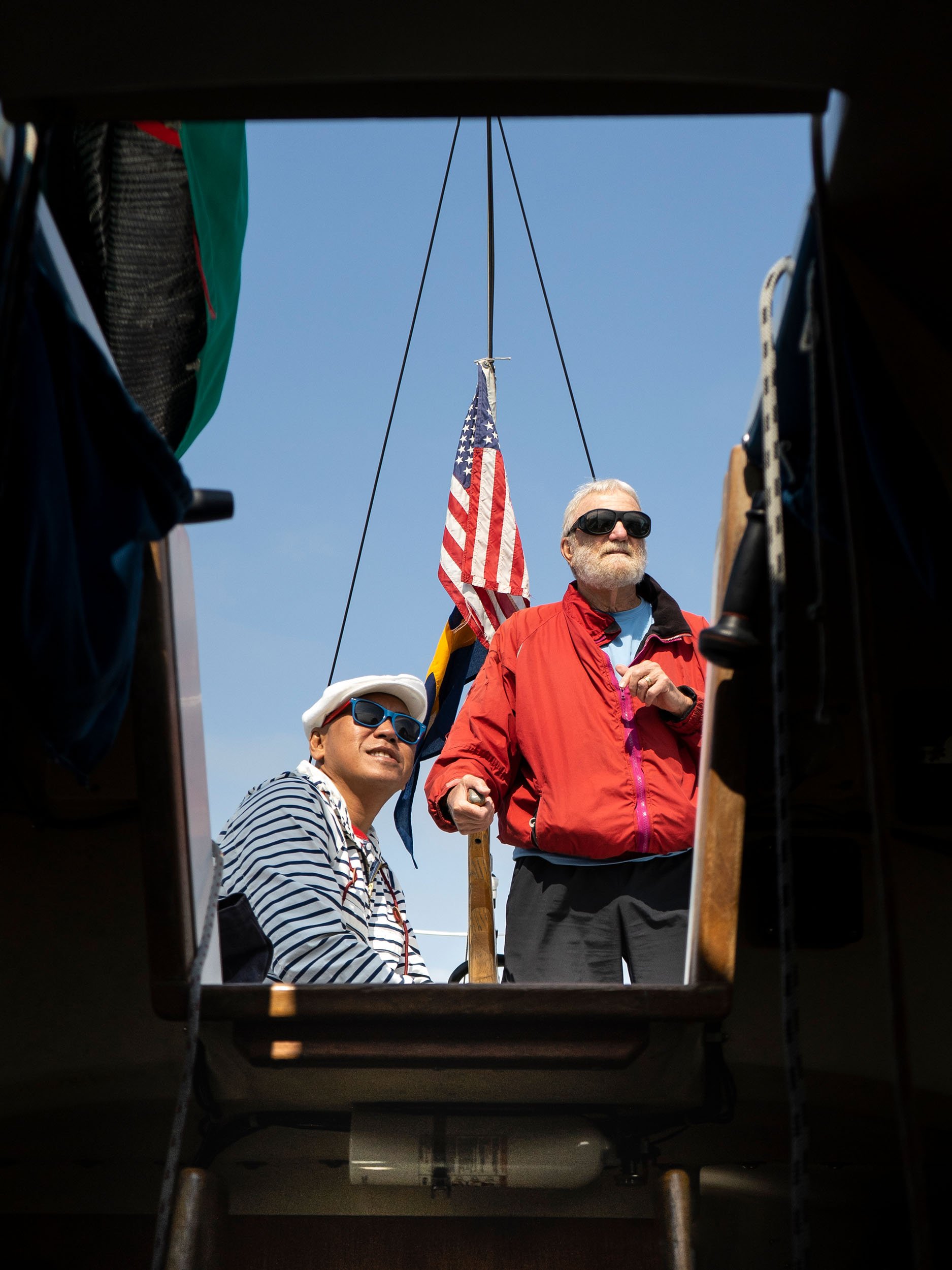 Two men standing outside on a sunny day, one holding an American flag, viewed from an opening in a dark structure.