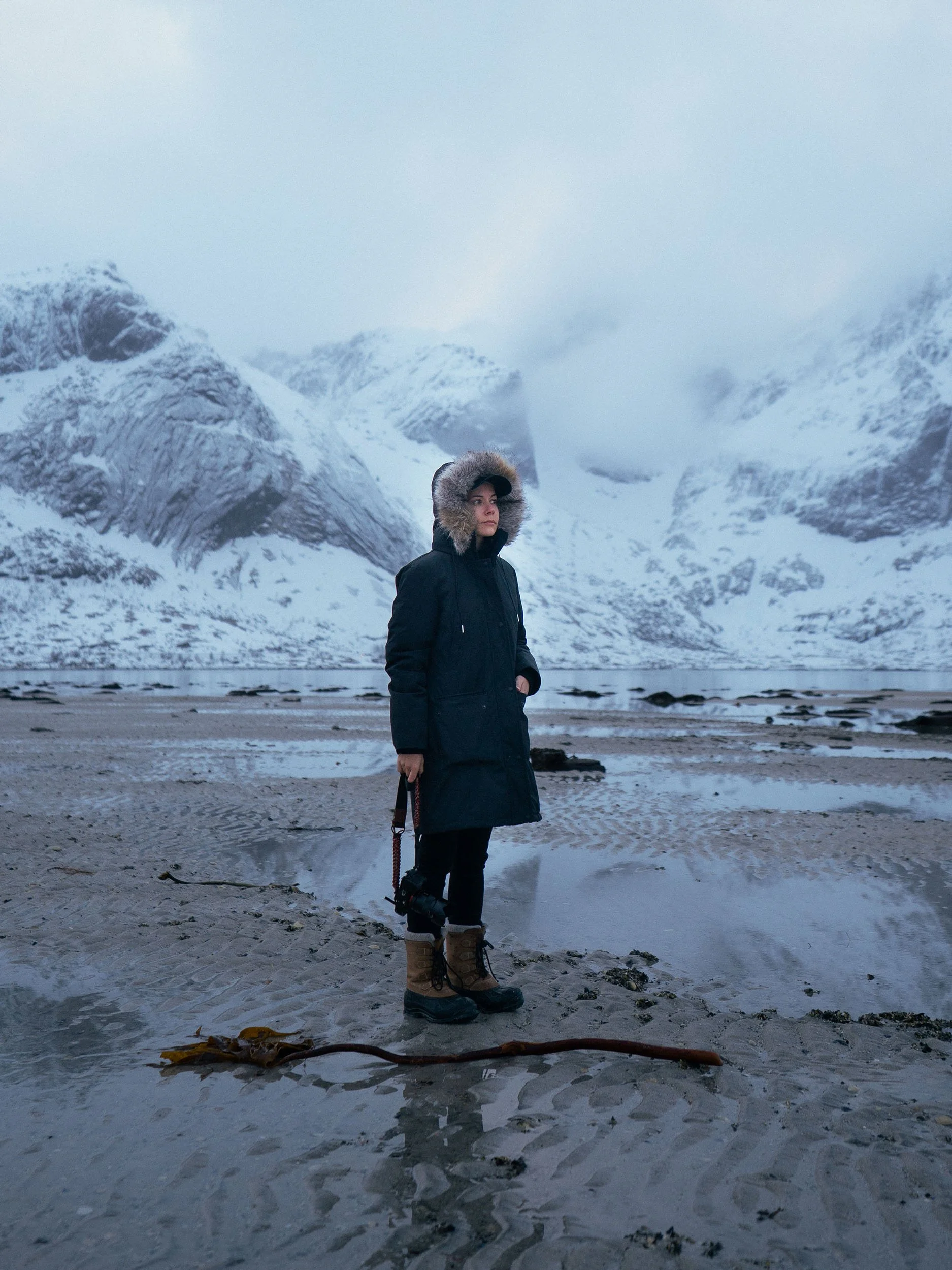 A woman wearing a black coat with a fur-lined hood and brown winter boots standing on a sandy, wet beach with snow-covered mountains in the background.