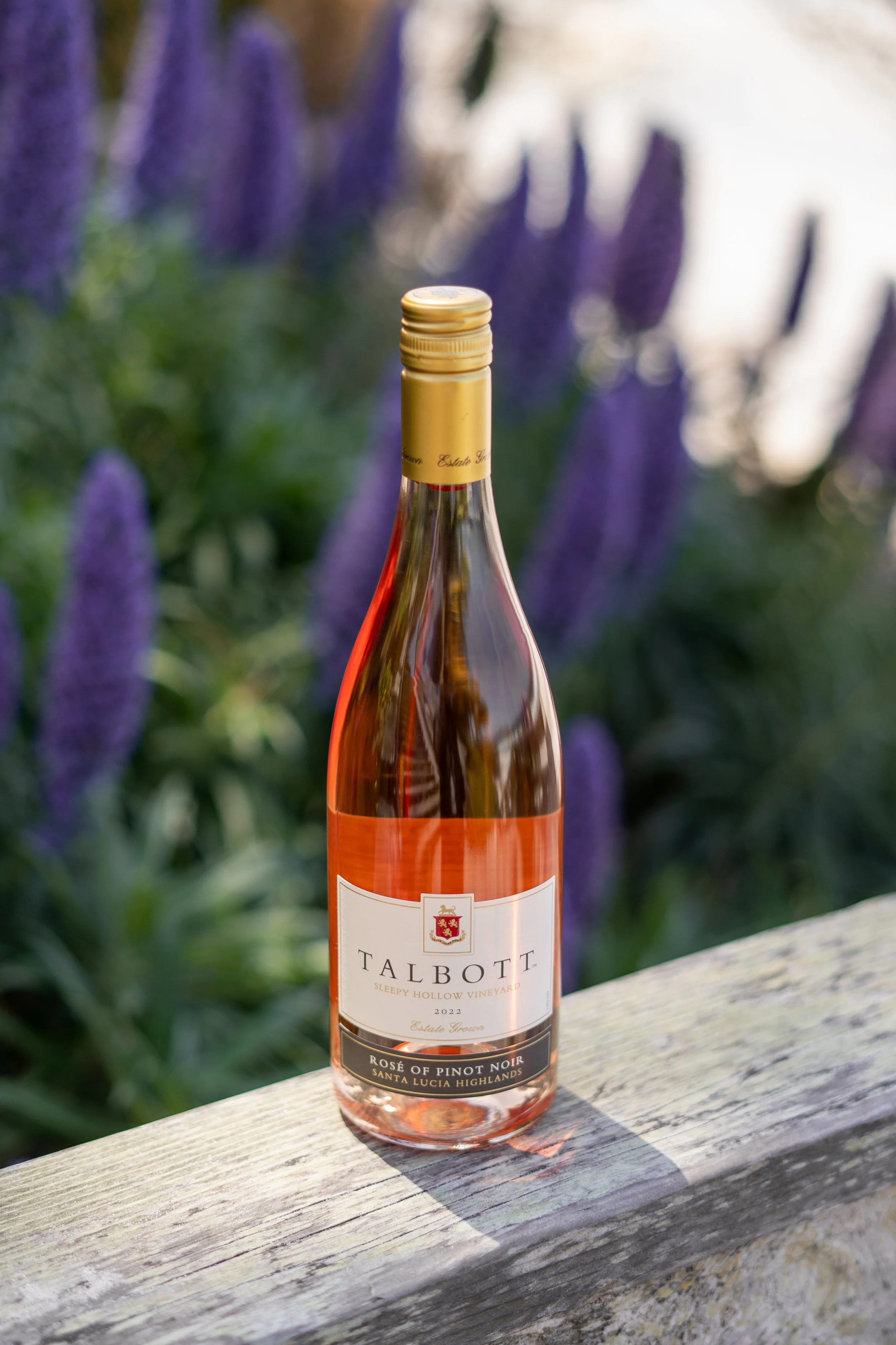 A bottle of Talbott Sleepy Hollow Vineyard Rosé of Pinot Noir wine resting on a weathered wooden surface outdoors with purple flowers and green foliage in the blurred background.