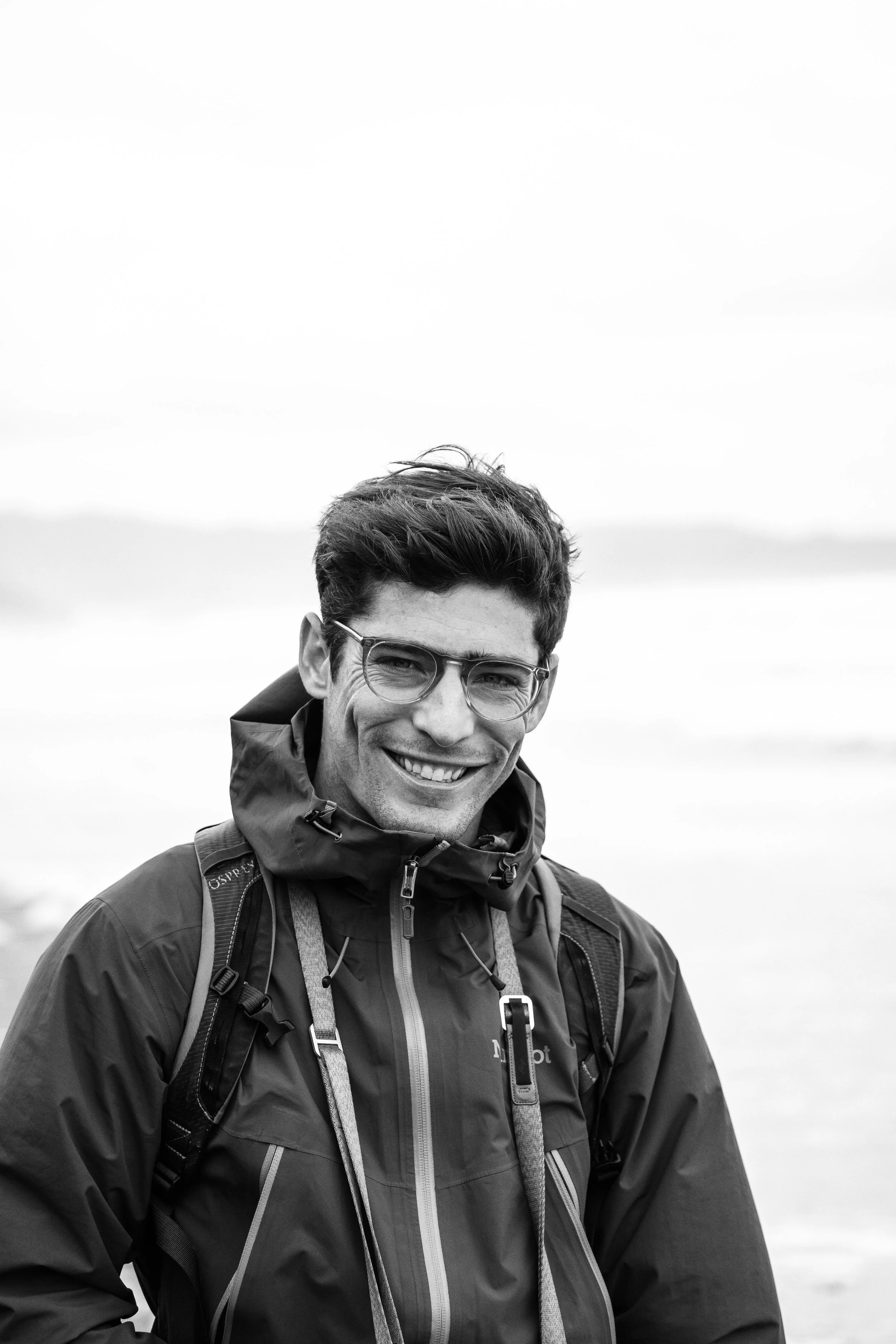 Black and white photo of a young man outdoors, smiling, wearing glasses and a hooded outdoor jacket with a backpack.