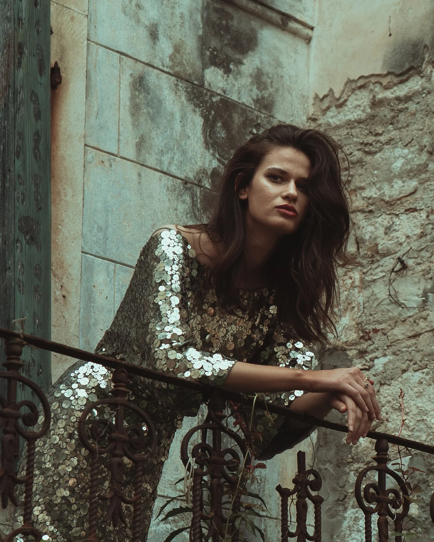A woman with long dark hair and makeup, wearing a sequined dress, sitting on a balcony with a wrought iron railing, leaning on her hands, against a weathered stone wall.