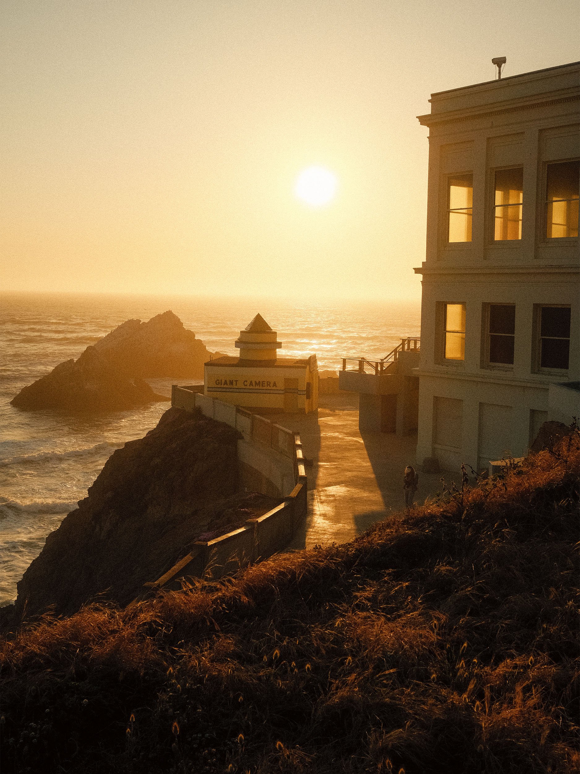 Sunset over the ocean with a building on the right side, a rocky formation in the water, and a sign that reads 'Giant Camera' in the foreground.