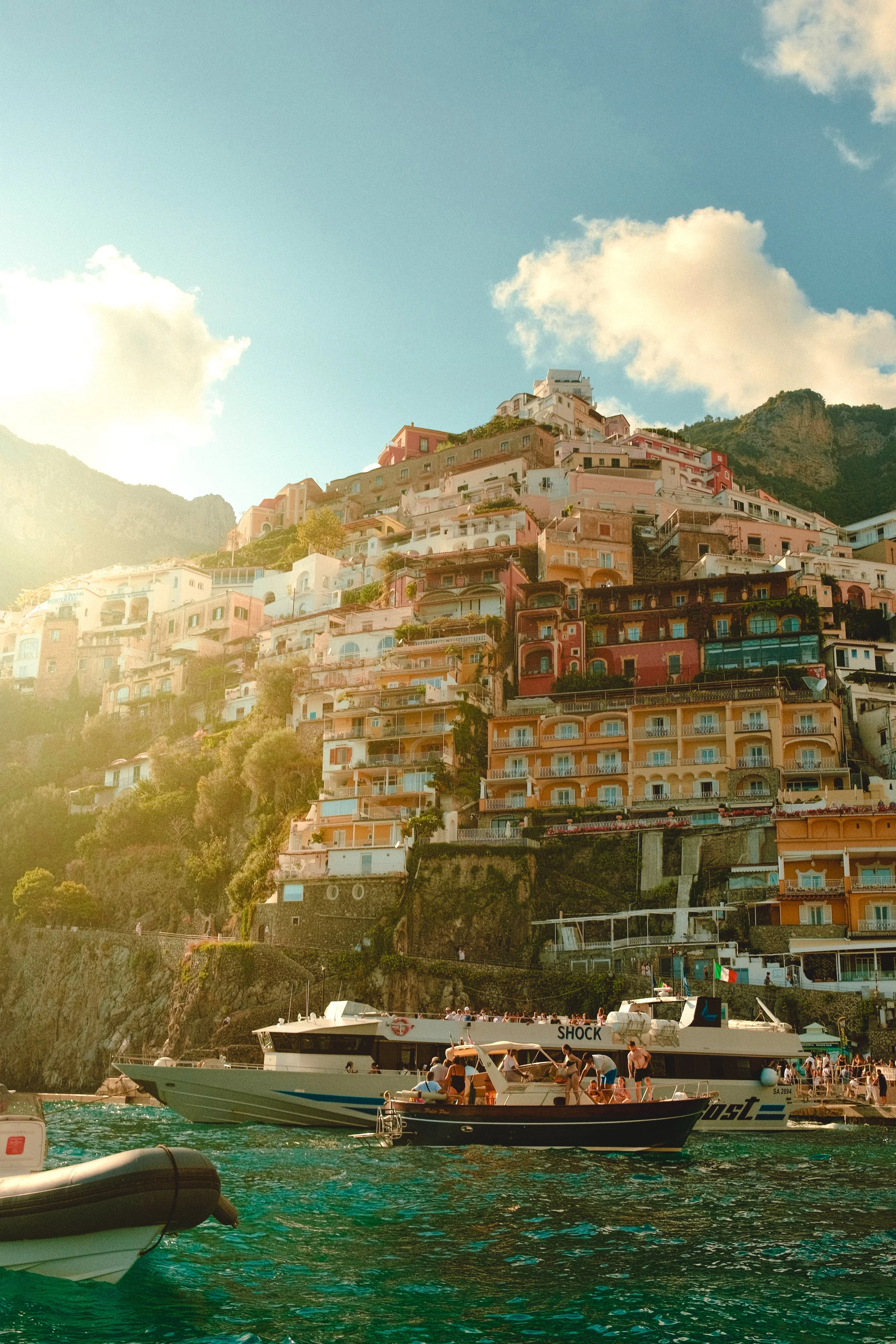 Colorful houses in Positano, Italy on a steep hillside overlooking the water, with boats docked below and a partly cloudy sky above.