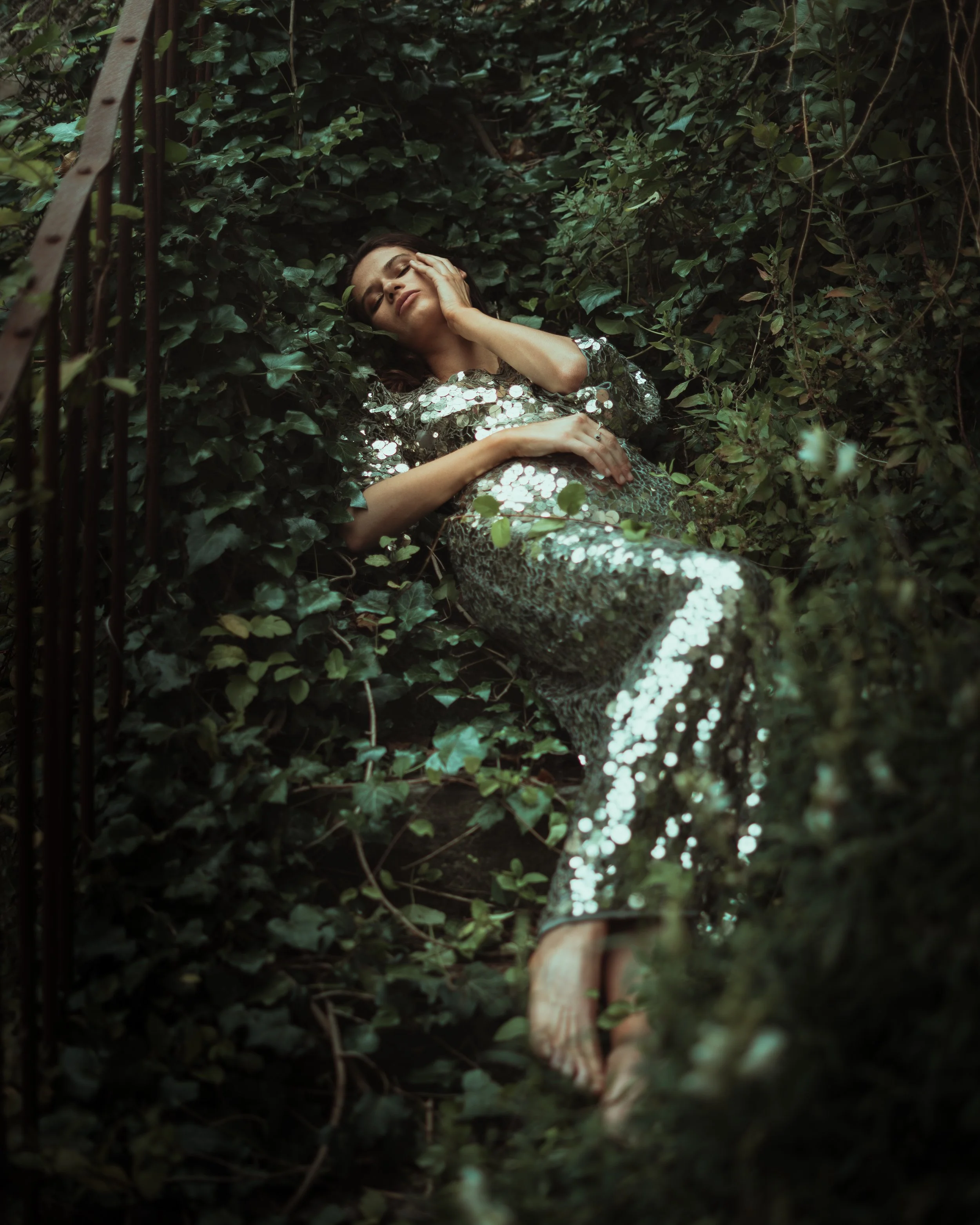 A woman lying amidst green foliage, wearing a shiny sequined dress, with her eyes closed and one hand resting on her face.