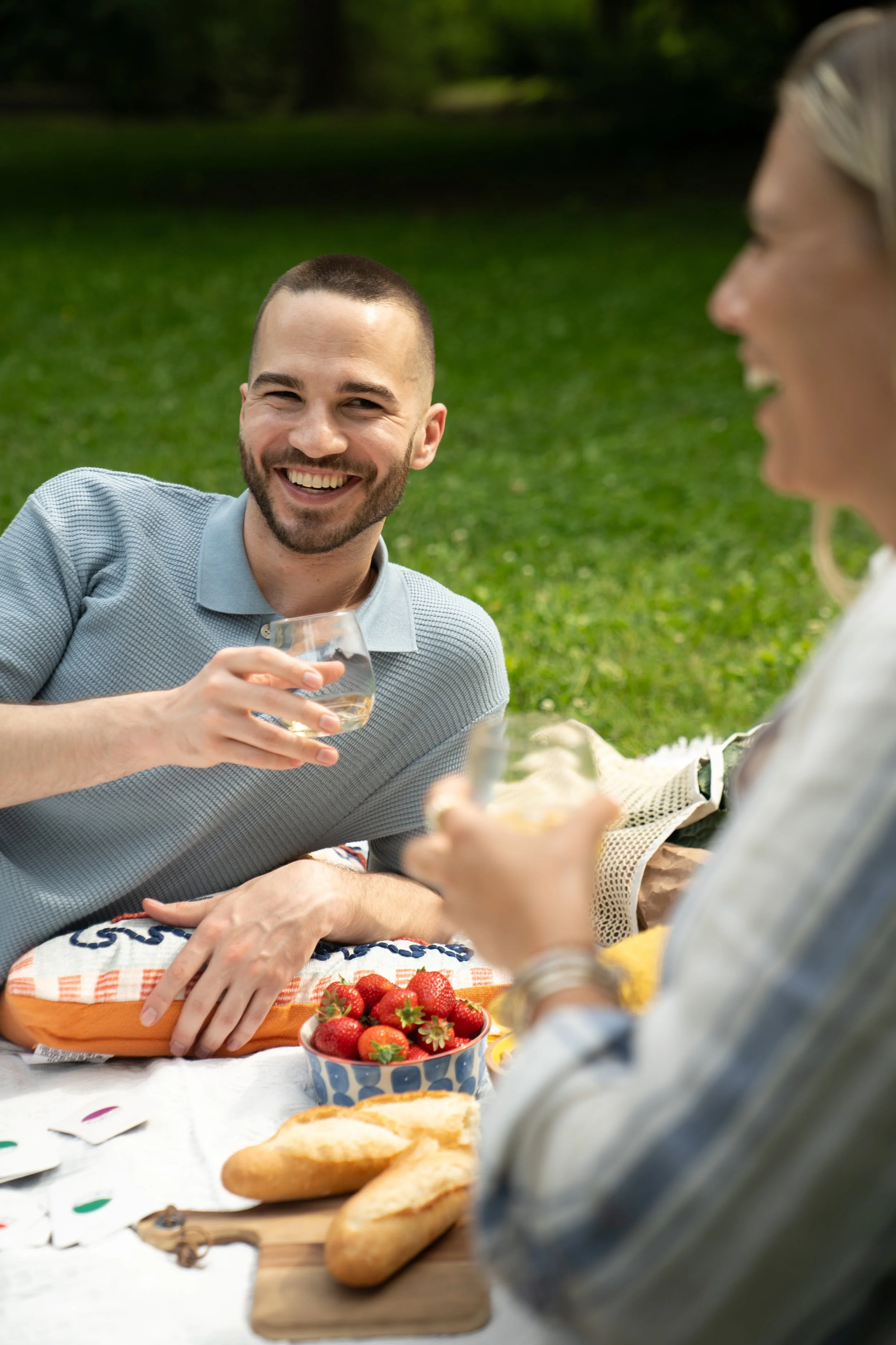 A man with short hair and a beard smiling and holding a glass of white wine, sitting outdoors on a grassy area with a woman partly visible in a light-colored top, also holding a glass, during a picnic with strawberries, bread, and other food on a table.