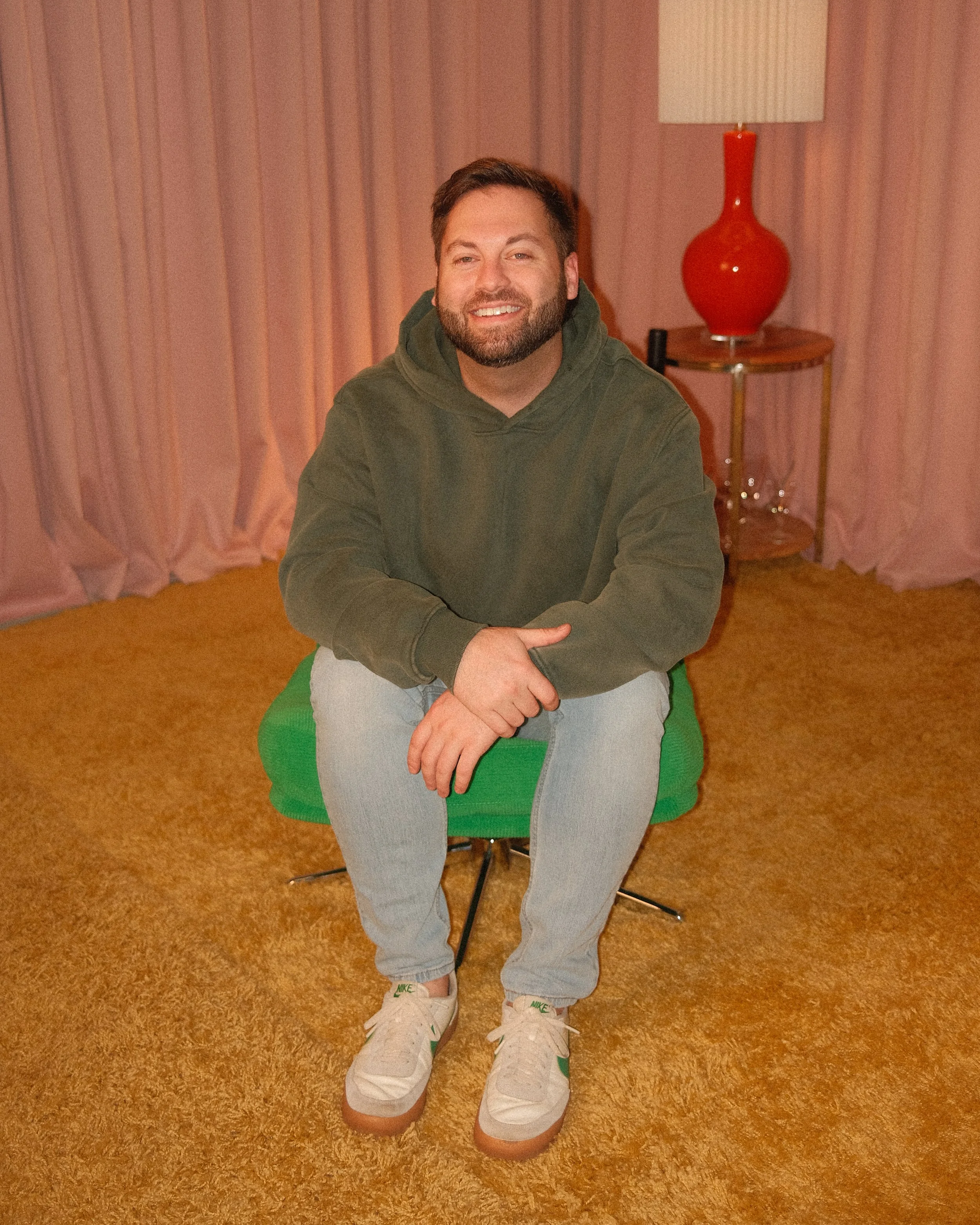 A man with a beard and short hair sitting on a green chair in a room with pinkish curtains and a yellow shag carpet, wearing a green hoodie, light blue jeans, and white sneakers with green accents, smiling at the camera.