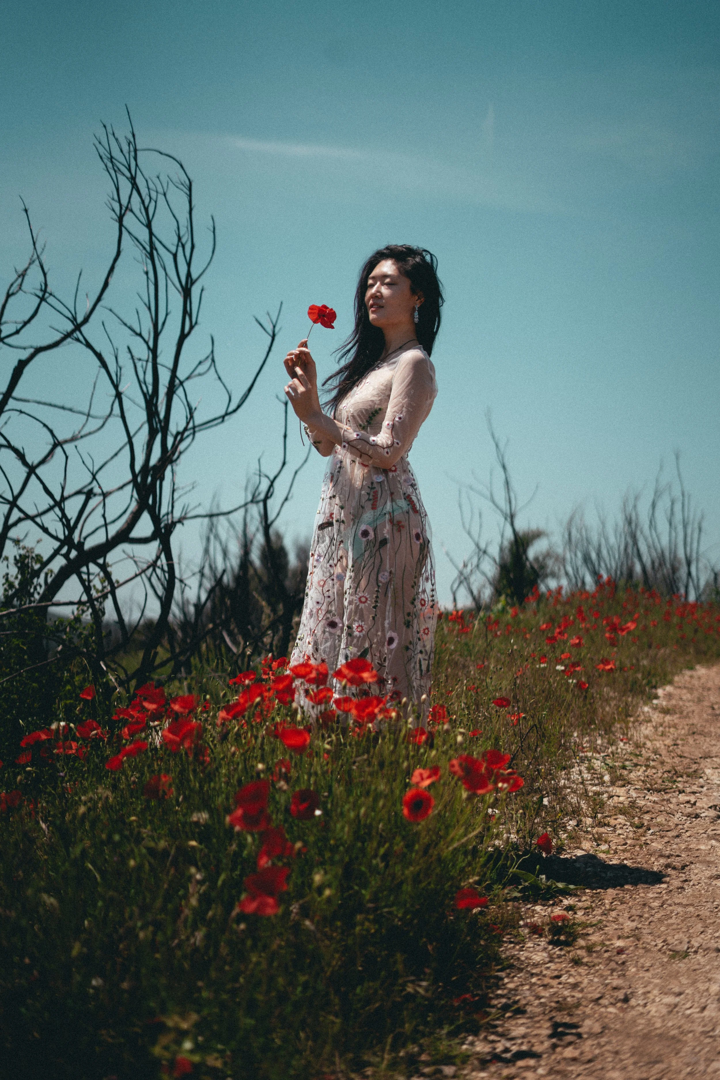 A woman in a long floral dress standing on a dirt path surrounded by red poppy flowers, holding a single red flower. She is outdoors on a clear day with a blue sky and some leafless branches nearby.