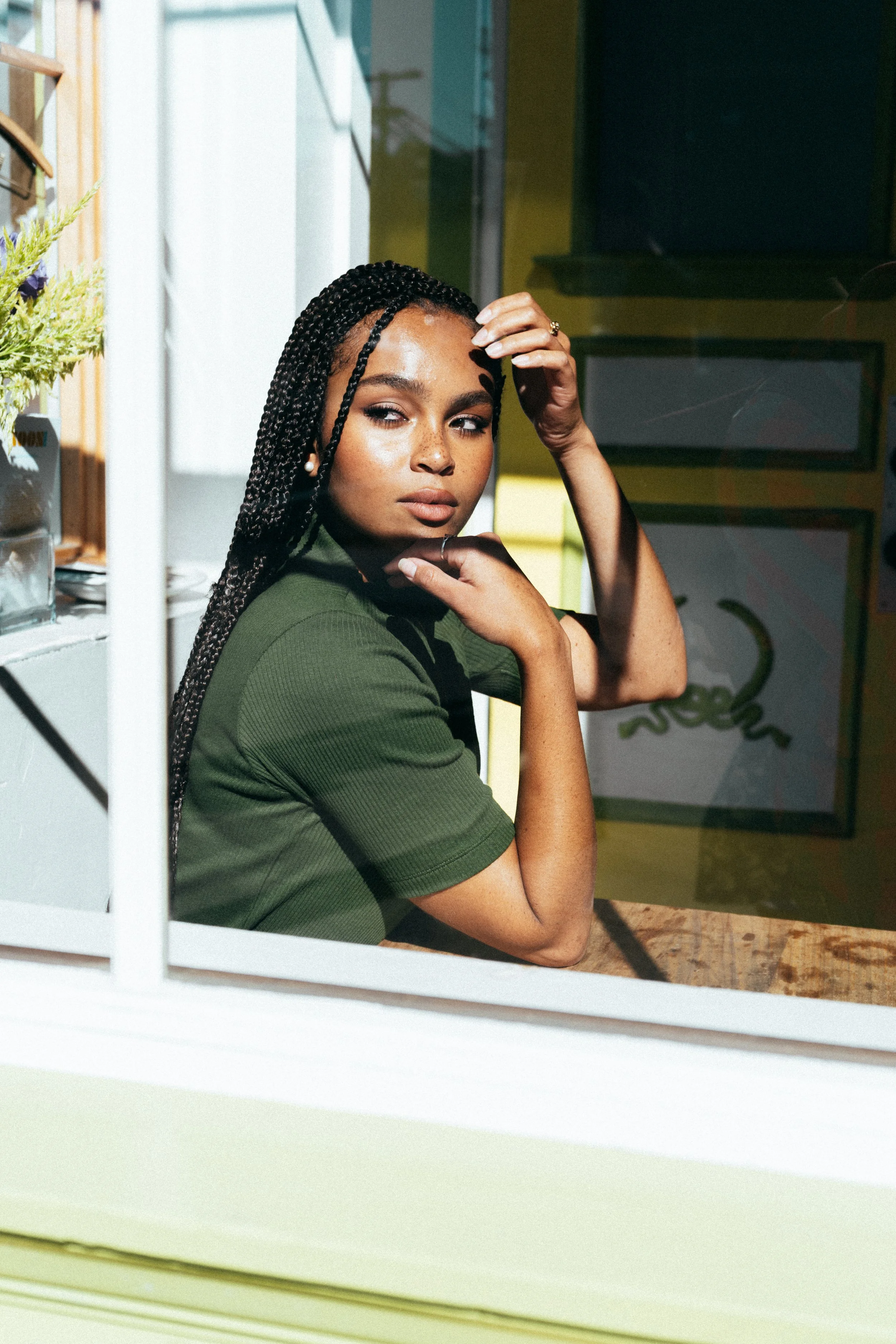 A woman with braided hair and a green shirt sitting at a wooden table, resting her arm on the table and touching her head, inside a room with bright natural light.