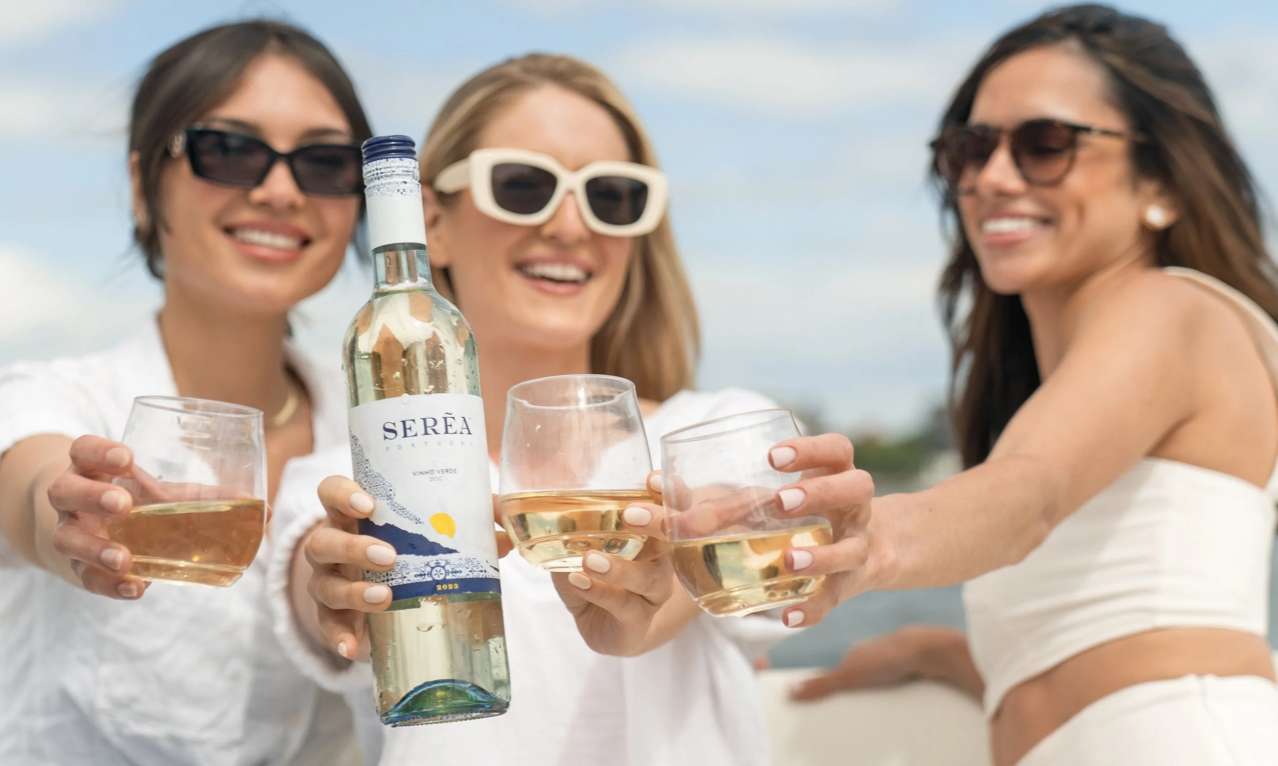 Three women smiling and holding glasses of white wine outdoors, with a blue sky in the background. One woman in the middle is holding a bottle of Serea Vinho Verde wine.