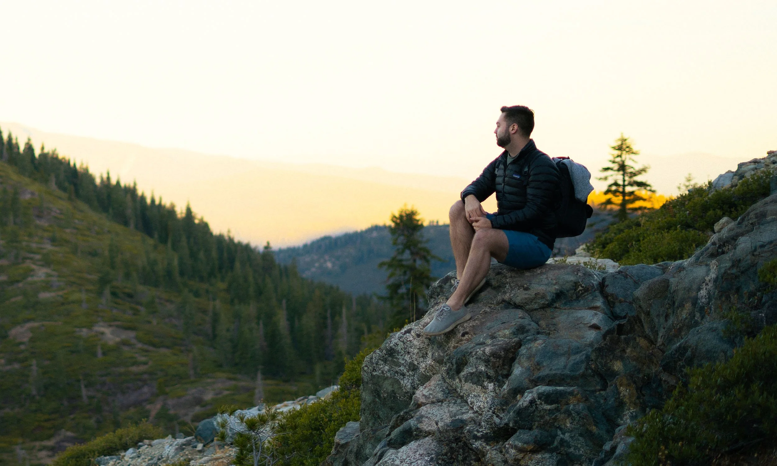 A man sitting on a rocky ledge overlooking a mountain valley at sunset.