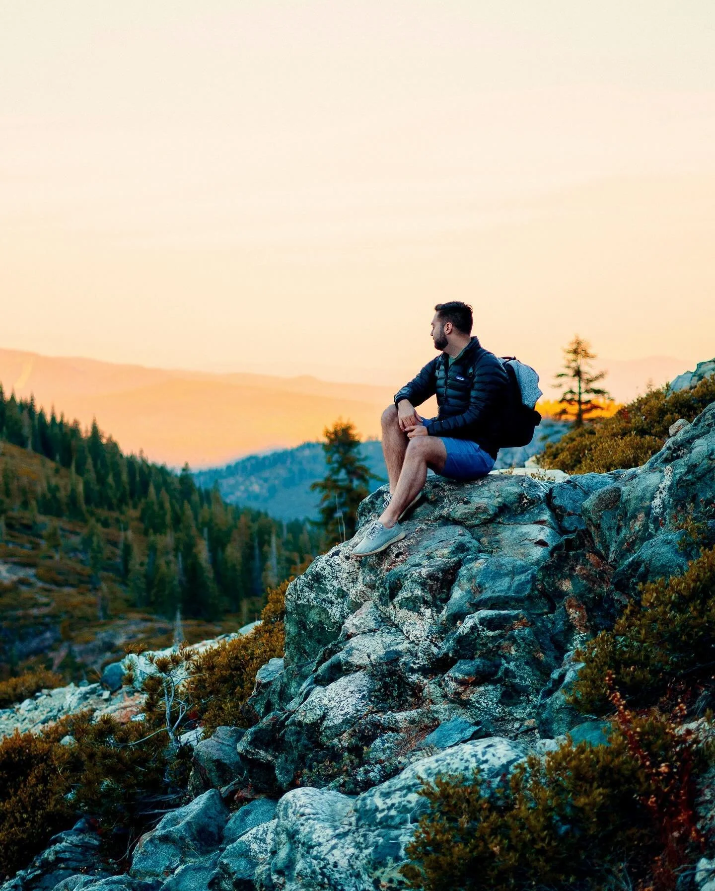 A man sitting on a large rock overlooking a mountainous landscape at sunset, wearing a black jacket, shorts, and a backpack.