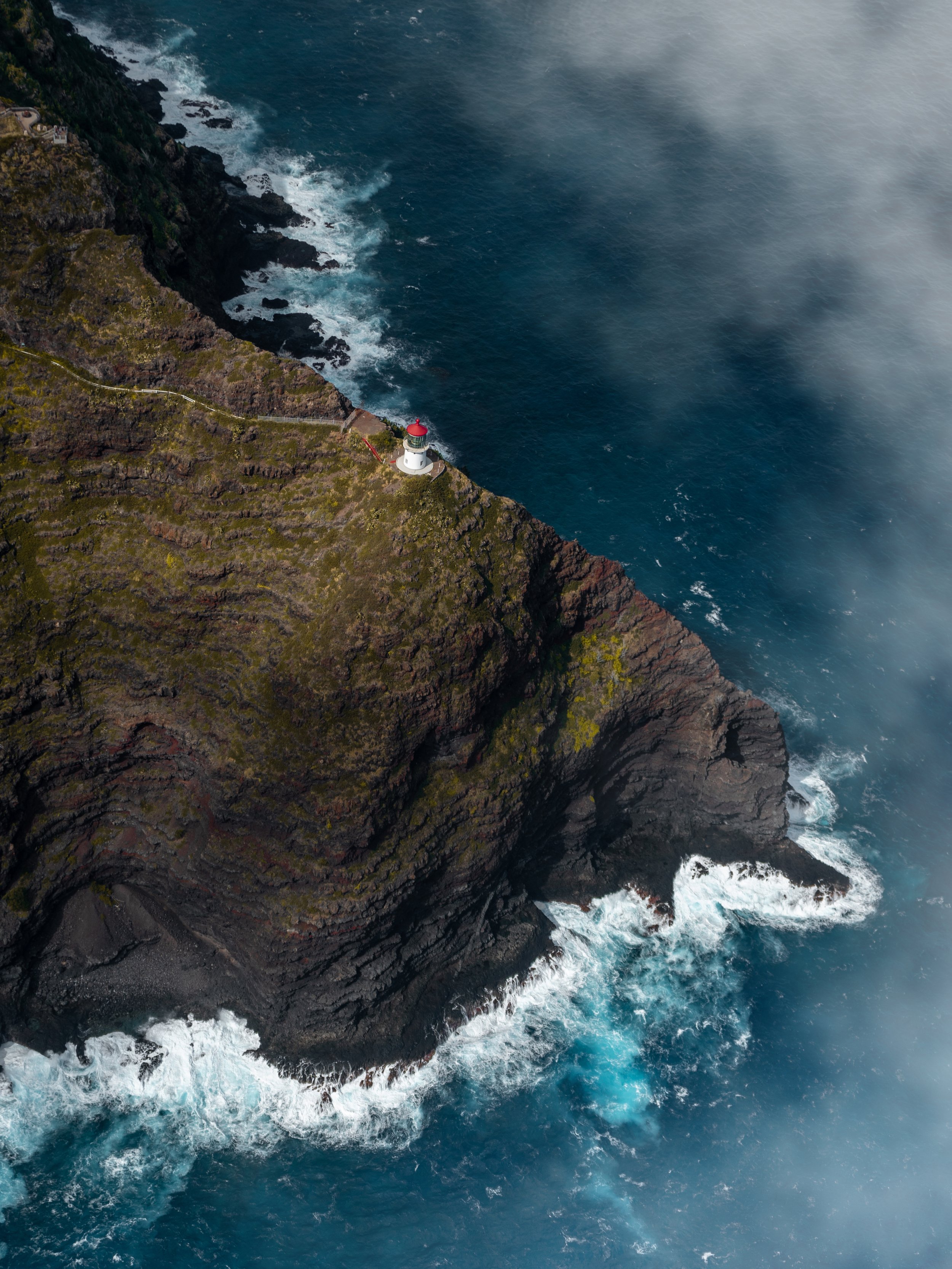 Aerial view of a rugged coastline with a lighthouse on a cliff overlooking the rough ocean, with waves crashing against the rocks.