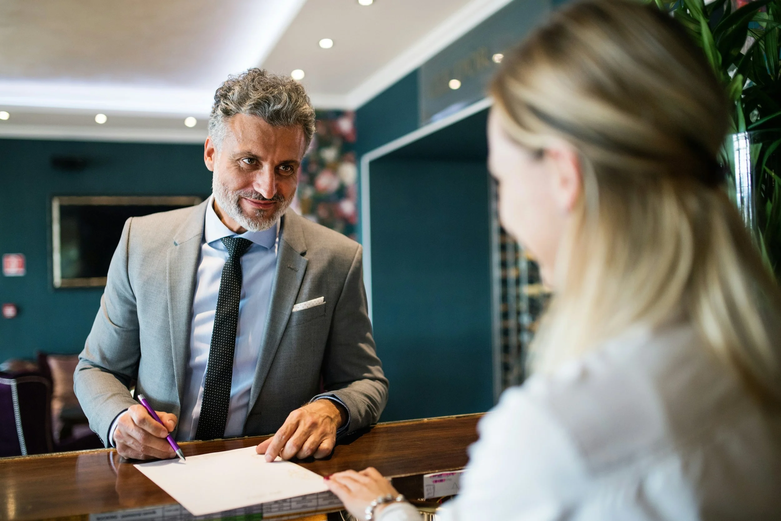 A man in a gray suit with a white shirt and dark tie handing a document to a woman at a reception desk in a hotel lobby.