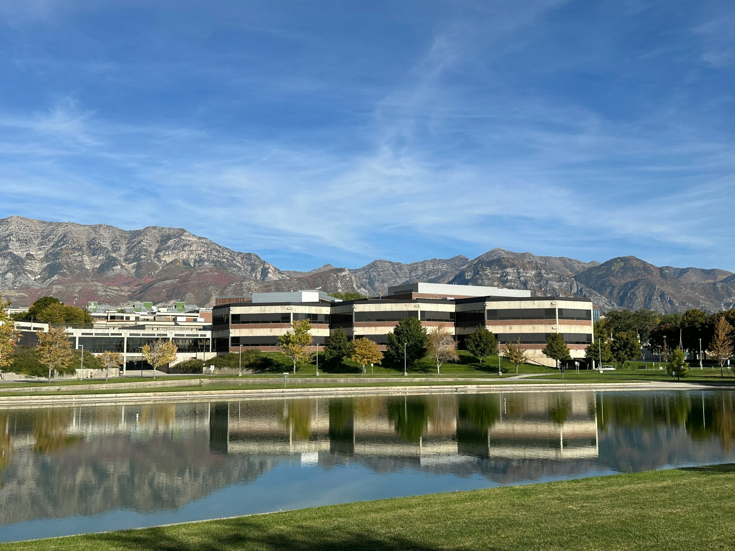 Modern office building with reflective windows, trees in front, a body of water reflecting the building and mountain range in the background under a partly cloudy blue sky.