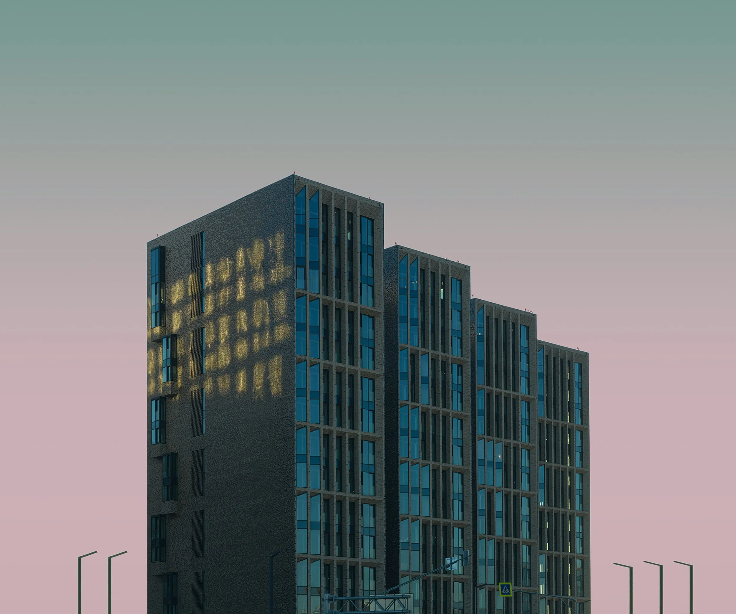 Modern multi-story building with large windows, reflected in the evening sky with pink and blue hues, street lamps and traffic signals in foreground.