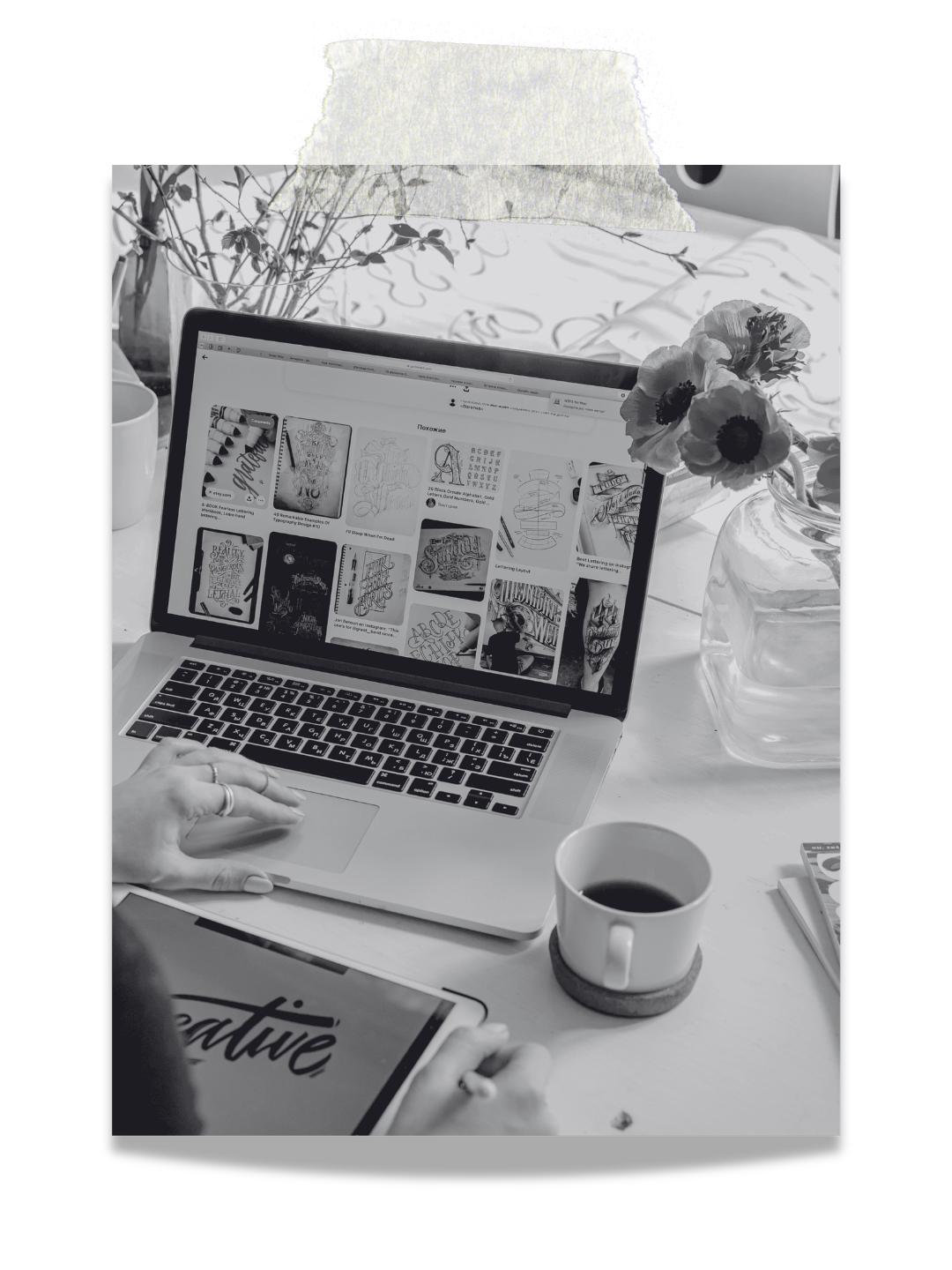 A workspace with a laptop displaying Pinterest-style images on a screen, a person's hand using a stylus or pen, a cup of coffee, a glass vase with flowers, a notebook, and a partially visible book or tablet on a white table.