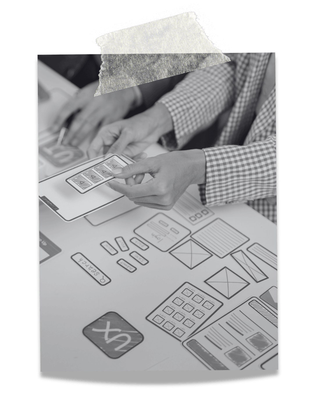 Black-and-white photo of two people working on a digital interface with various schematic diagrams, UI elements, and wireframes on a tablet and a table, indicating a design or planning session.