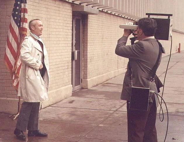A man in a trench coat posing for a photo next to an American flag, while another person takes his picture with a vintage camera setup on a sidewalk.