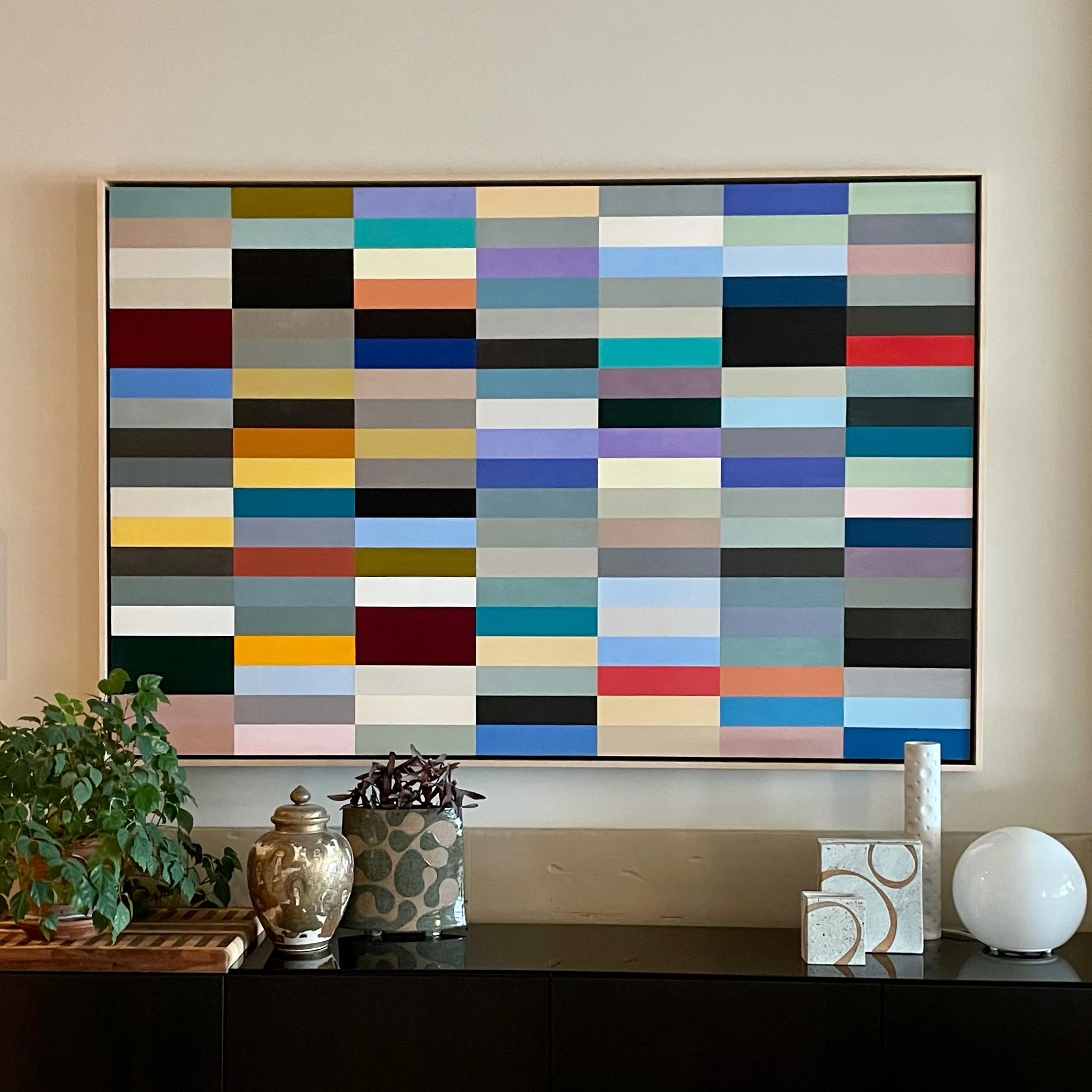 A large abstract artwork composed of many colorful rectangular blocks, hung on a beige wall above a black sideboard with decorative items including a potted plant, ceramic vases, and sculptures.