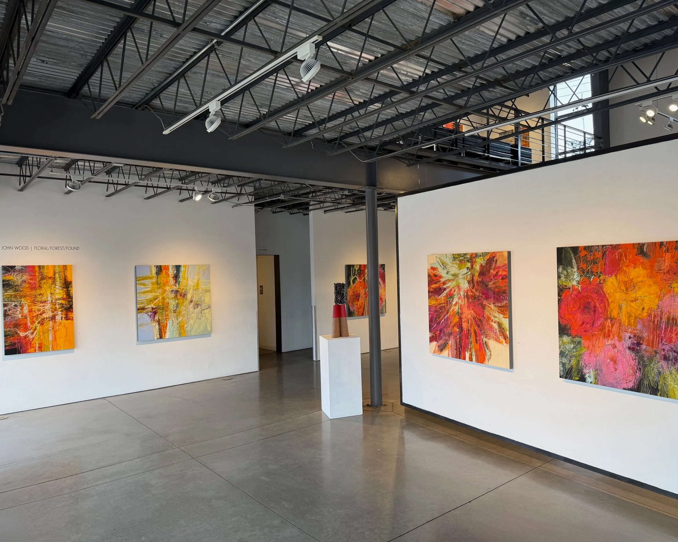 Interior of an art gallery with white walls displaying colorful abstract paintings, concrete floor, industrial ceiling with exposed beams and track lighting.