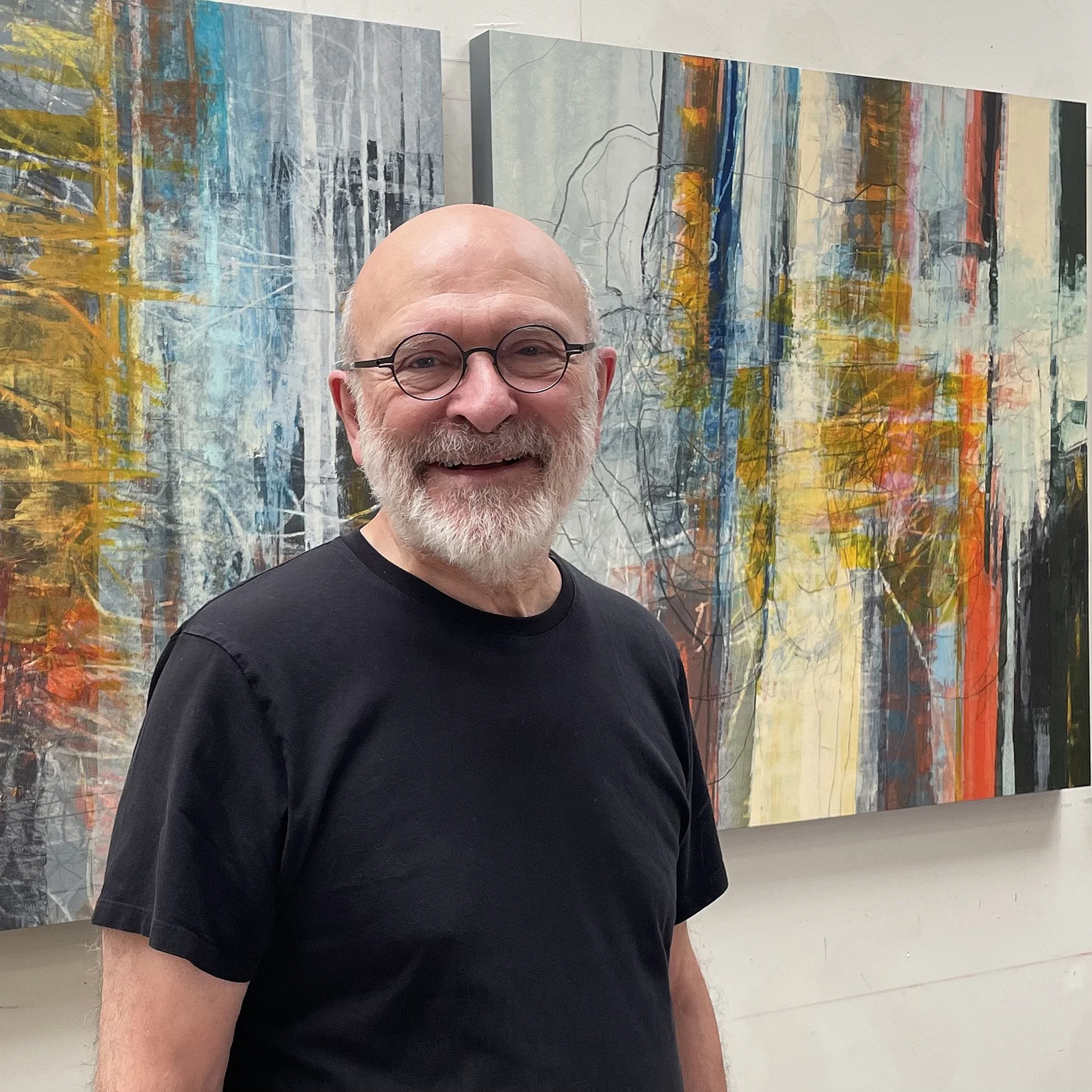 A smiling older man with glasses and a beard standing in front of abstract colorful paintings in an art gallery.