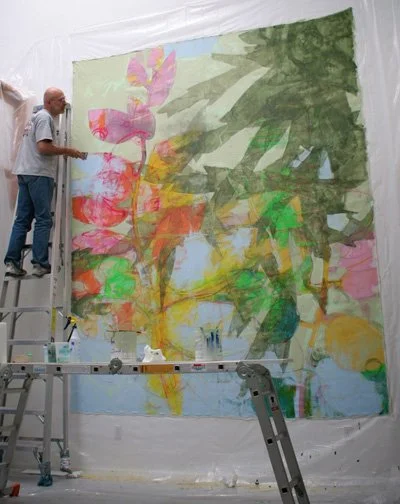 An artist is working on a large abstract painting of a plant with pink, yellow, green, and orange hues, set up in an art studio.