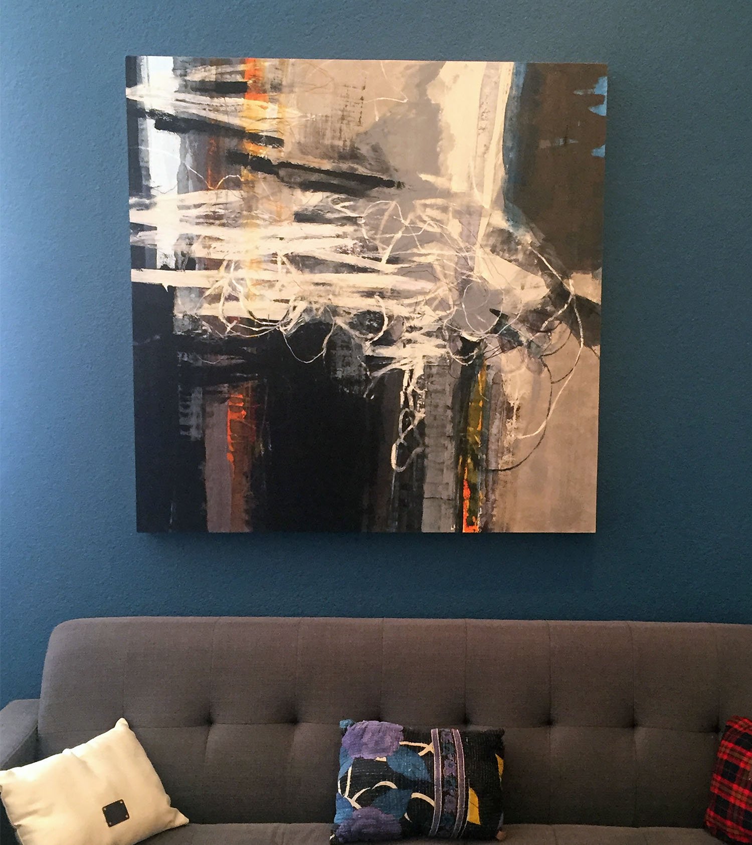 Abstract landscape painting hanging on dark teal wall above a gray couch with decorative pillows.