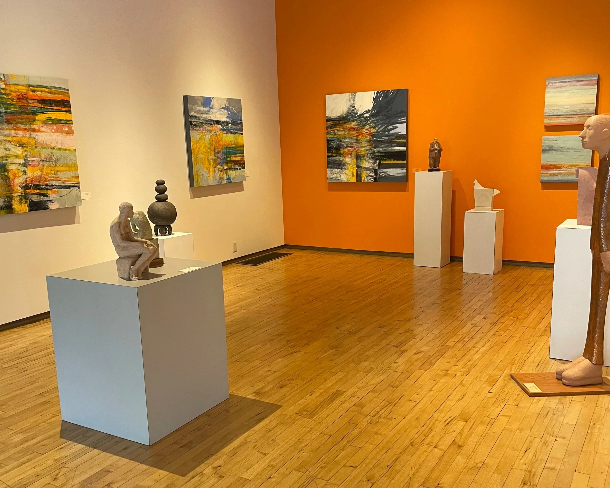 An art gallery with abstract paintings on the wall and sculptures on white pedestals, including a seated figure sculpture, a stacked stone sculpture, and a stylized head sculpture, all displayed on a wooden floor.