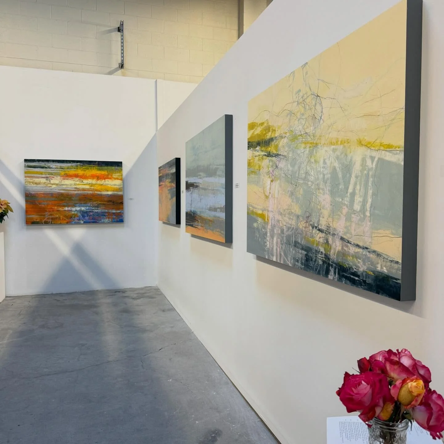 Art gallery with four abstract landscape paintings on a white wall, and pink flowers in a glass jar in the foreground.
