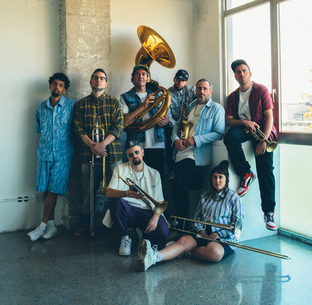 Grupo de ocho personas con instrumentos musicales en un interior junto a una ventana y una pared de concreto.