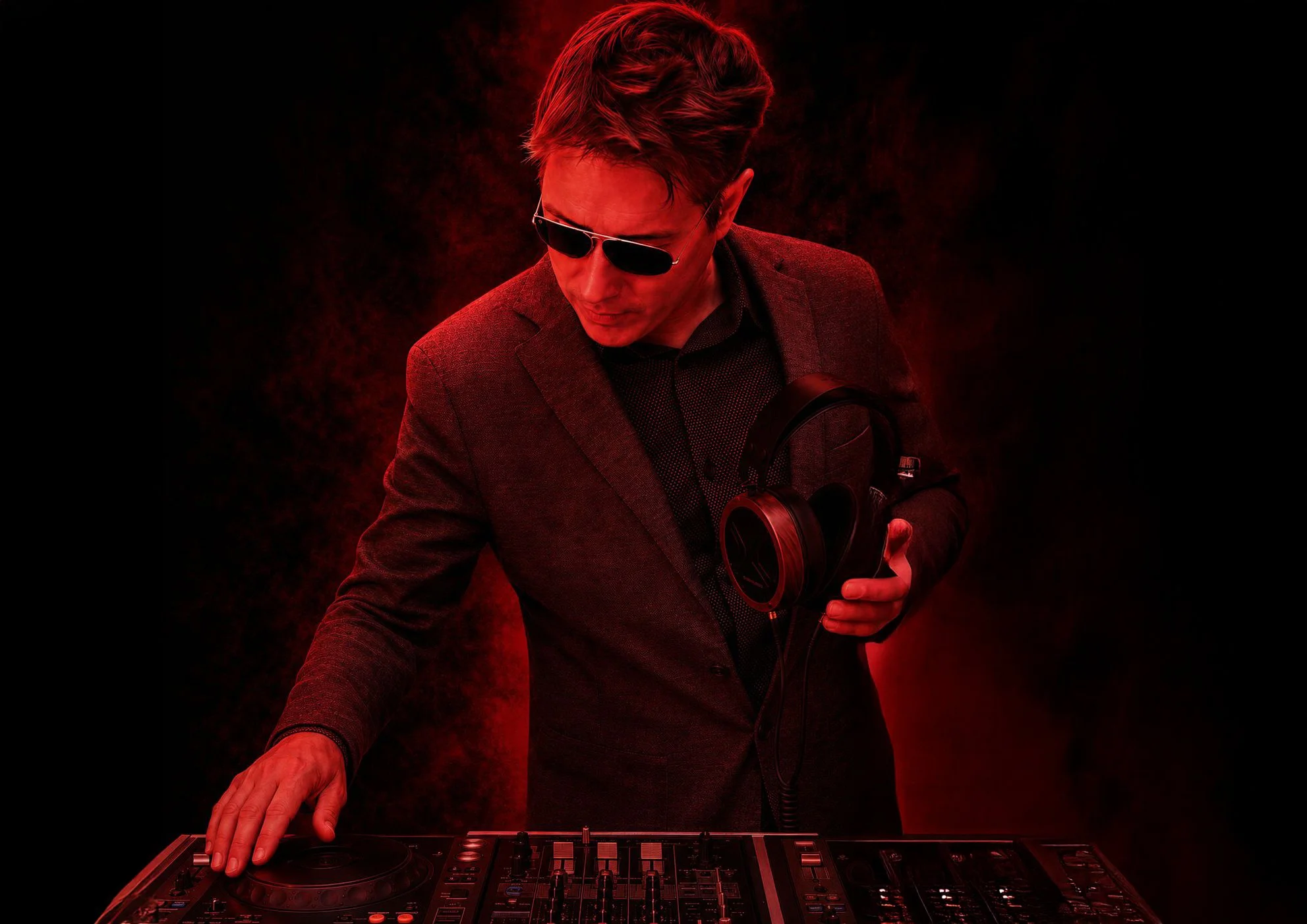 Hombre con gafas de sol y chaqueta ajustada, tocando una consola de DJ en un fondo oscuro con tonos rojos.