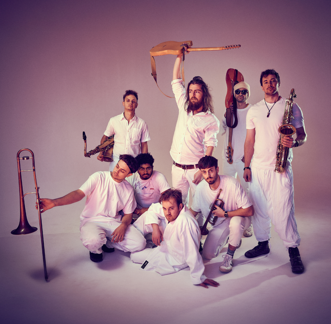 Grupo de siete hombres con instrumentos musicales, algunos con ropa blanca y otros con accesorios, en un fondo neutro, en una pose expresiva.