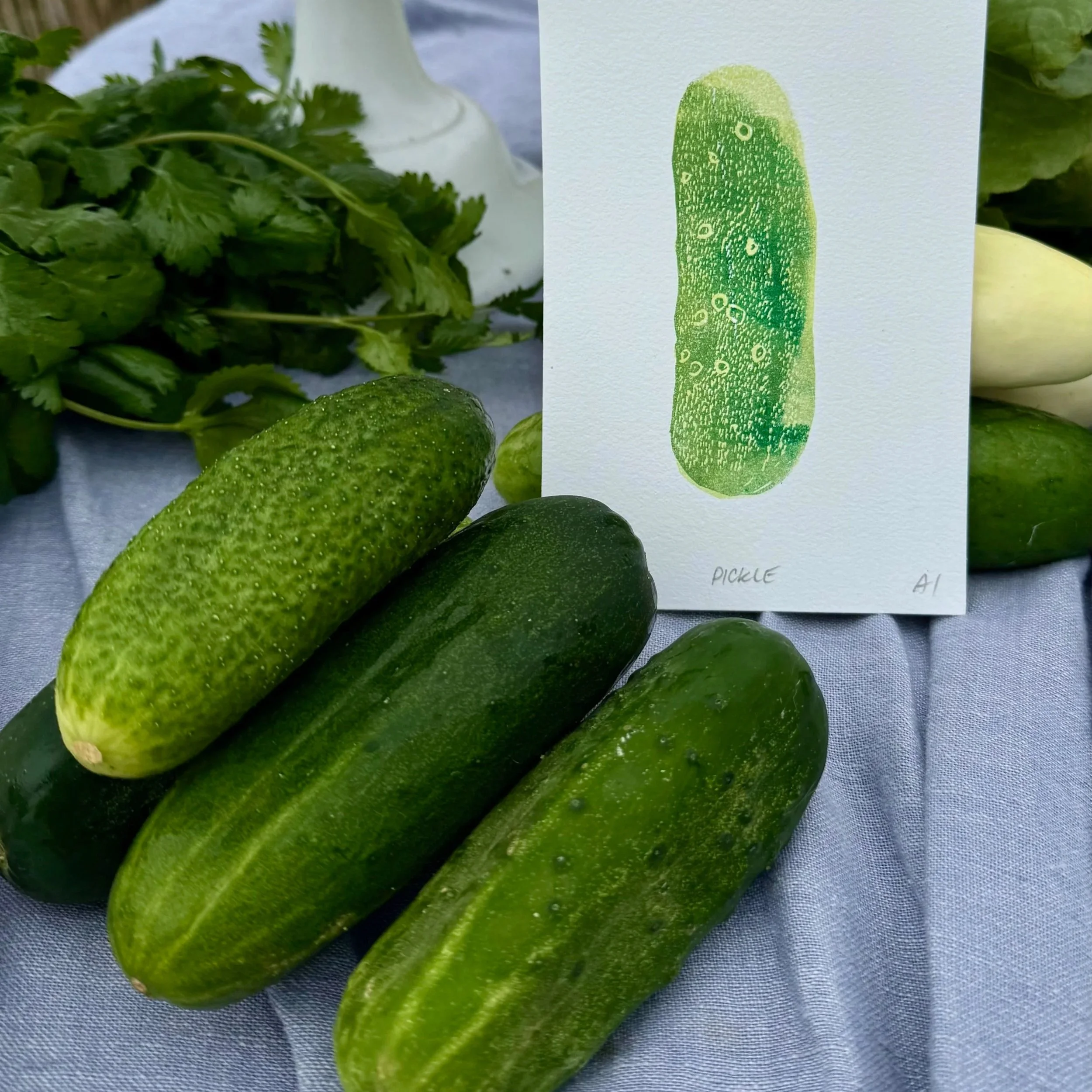 Three cucumbers and a bunch of cilantro on a table, with a card featuring a stylized cucumber illustration labeled 'PICKLE'.