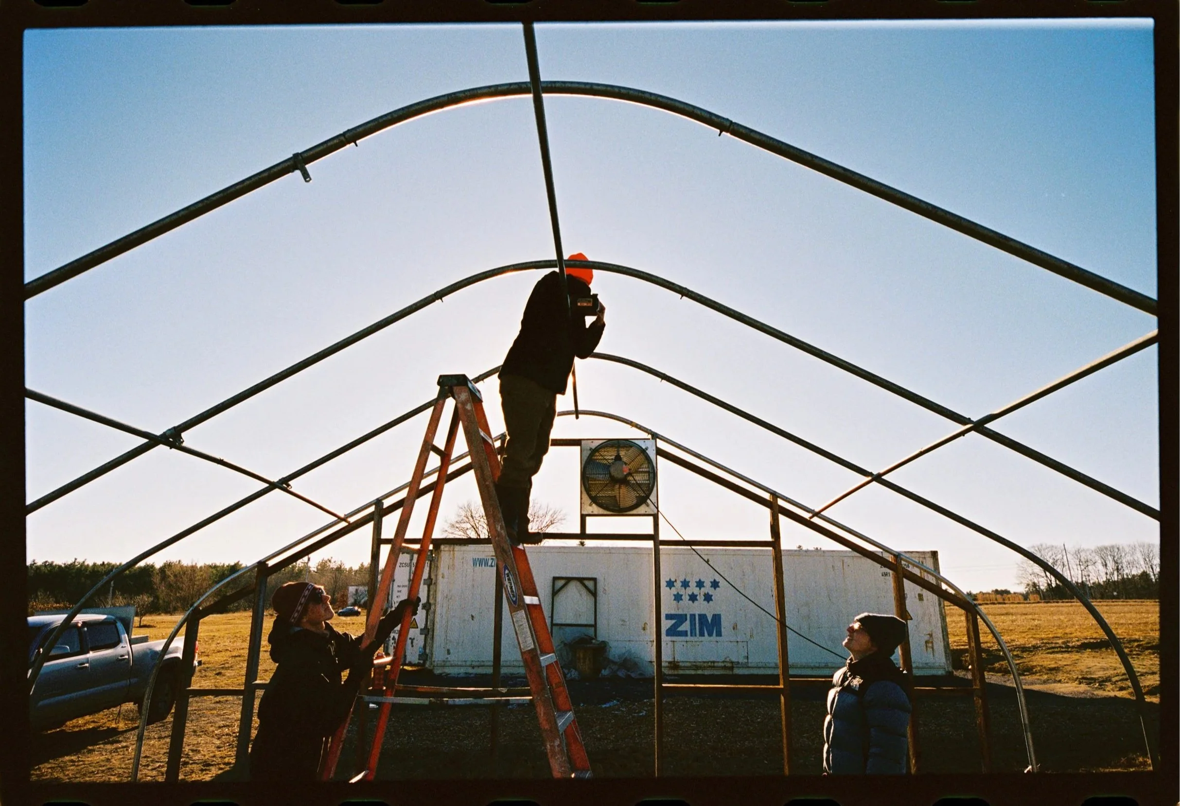 Three people working on the metal frame of a greenhouse structure outdoors during daytime, one on a ladder and two on the ground, with a pickup truck and open field in the background.