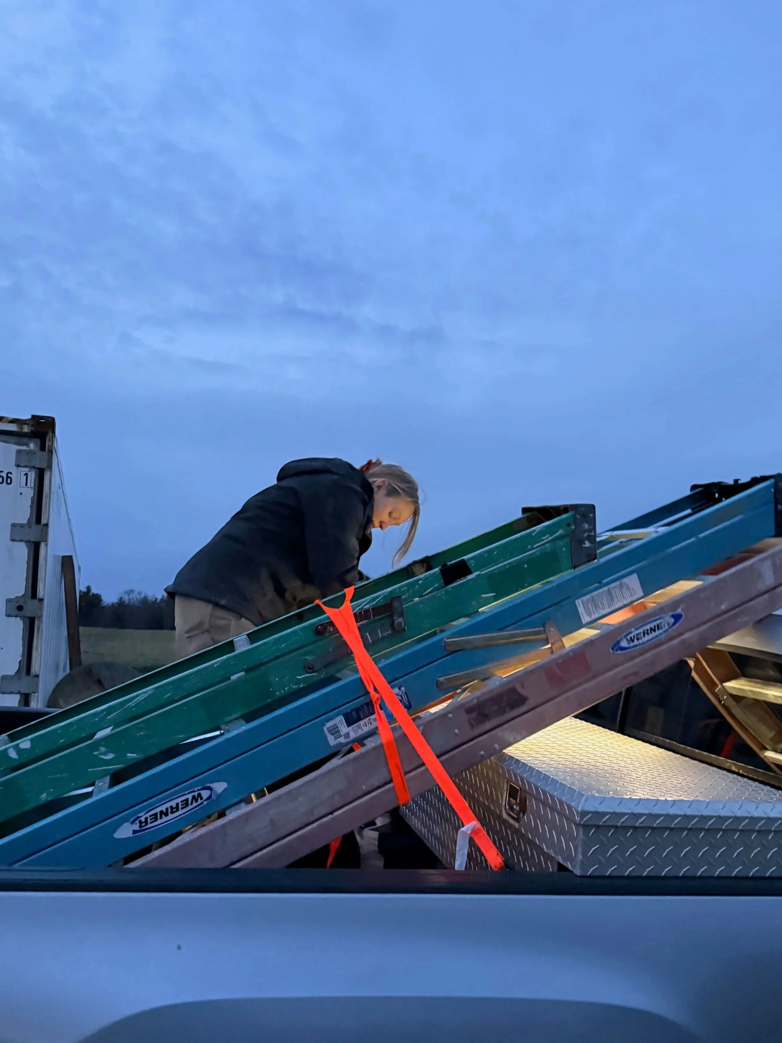 A woman is loading or unloading ladders and equipment onto a truck bed during dusk or dawn in an outdoor setting.