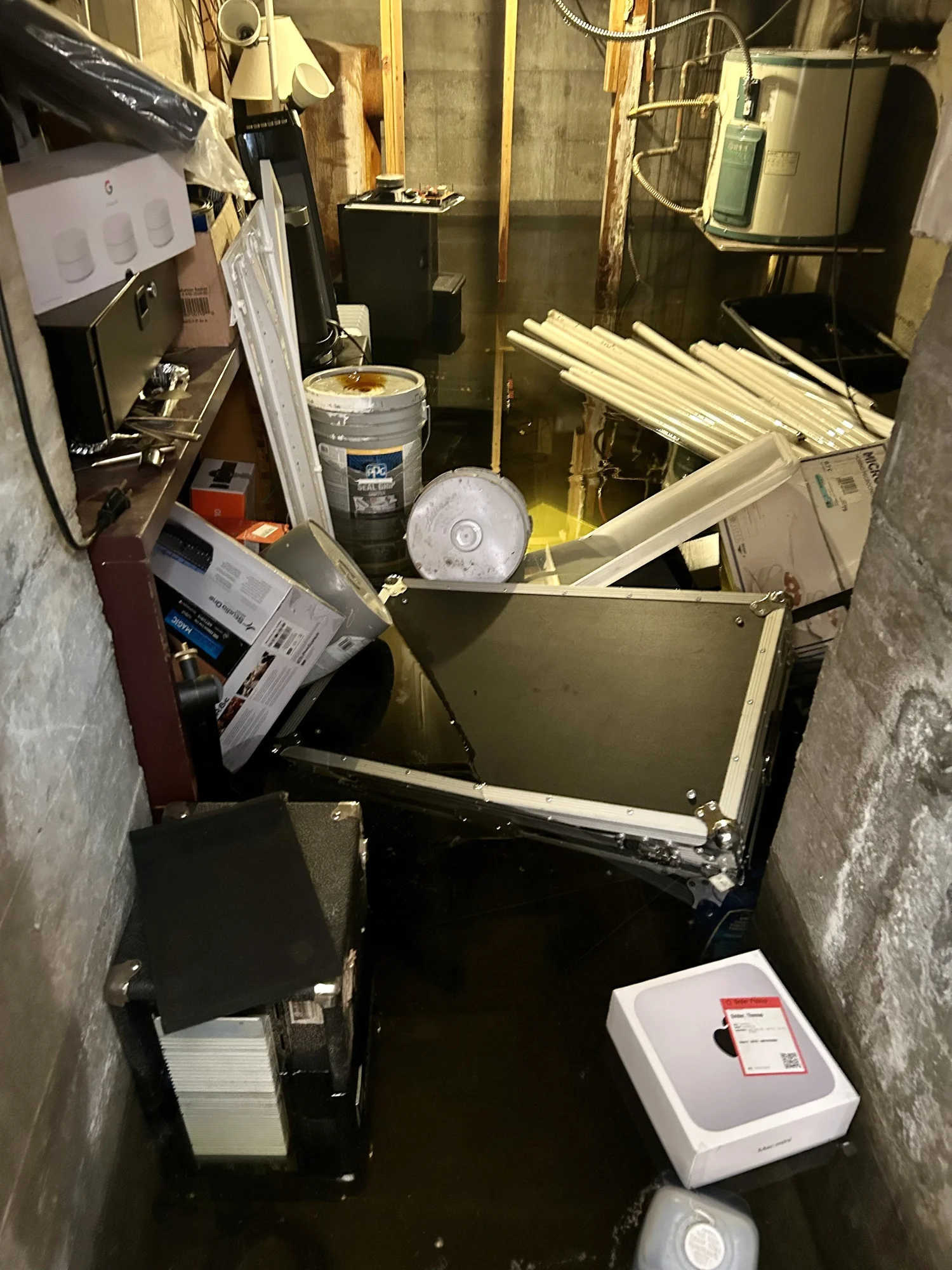 Flooded basement with water covering the floor, a fallen ceiling tile, pipes, boxes, and equipment.