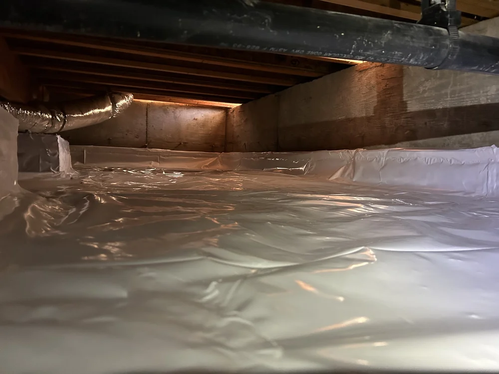 Attic space with wooden beams and insulation covered with clear plastic sheeting.