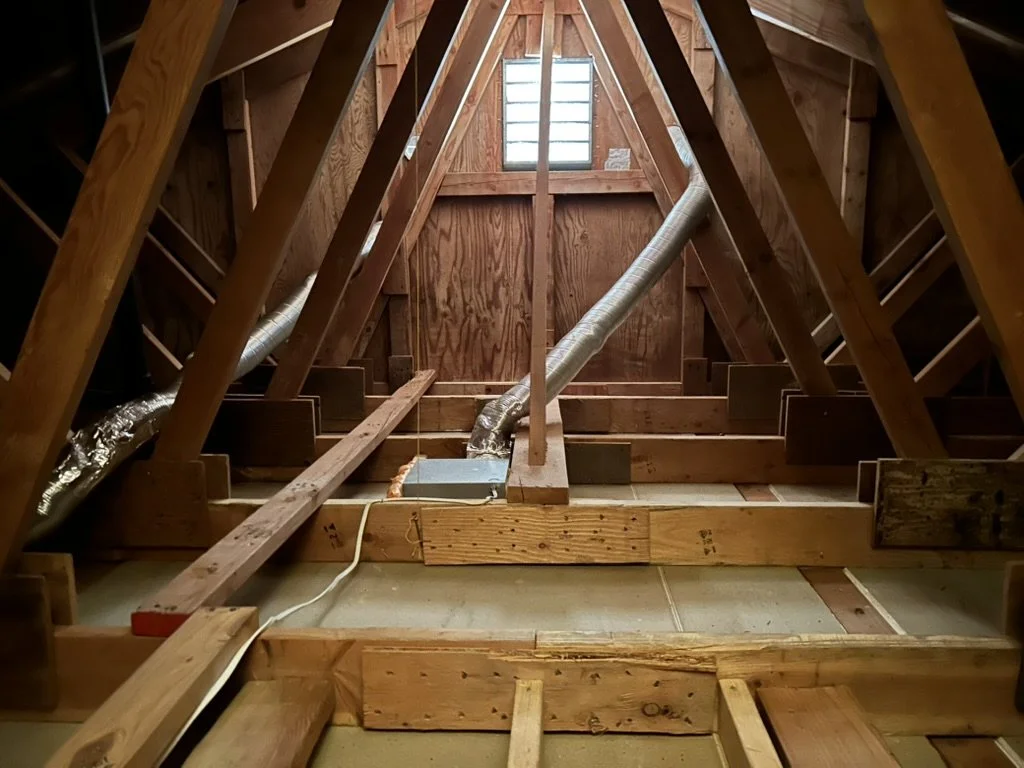 View of an attic with wooden framing, insulation ducts, and a small vent window.