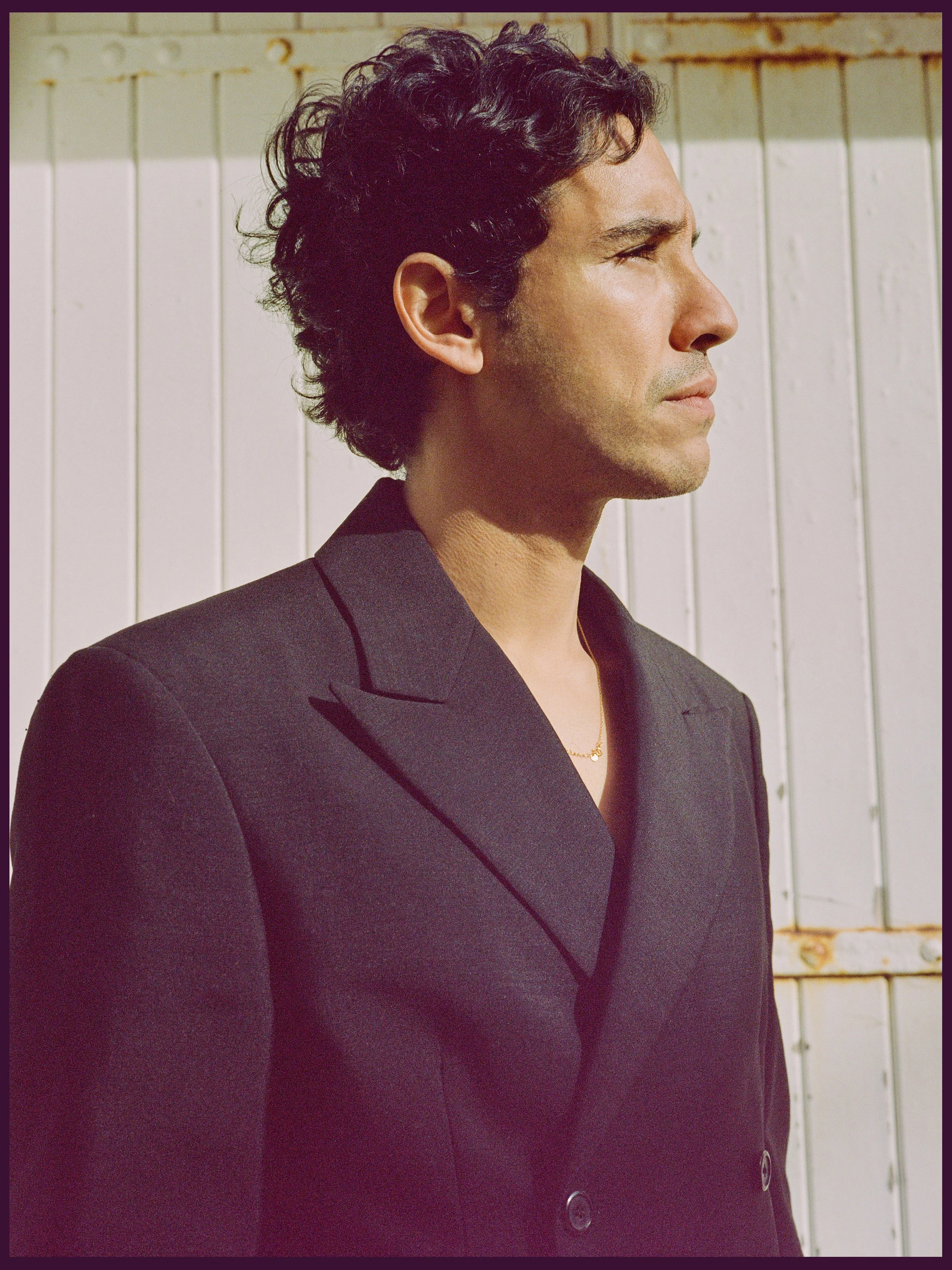 Profil d'Adam Bessa portant un blazer noir foncé avec des boutons, fermée, avec une chaîne en or, devant un mur en bois blanc.