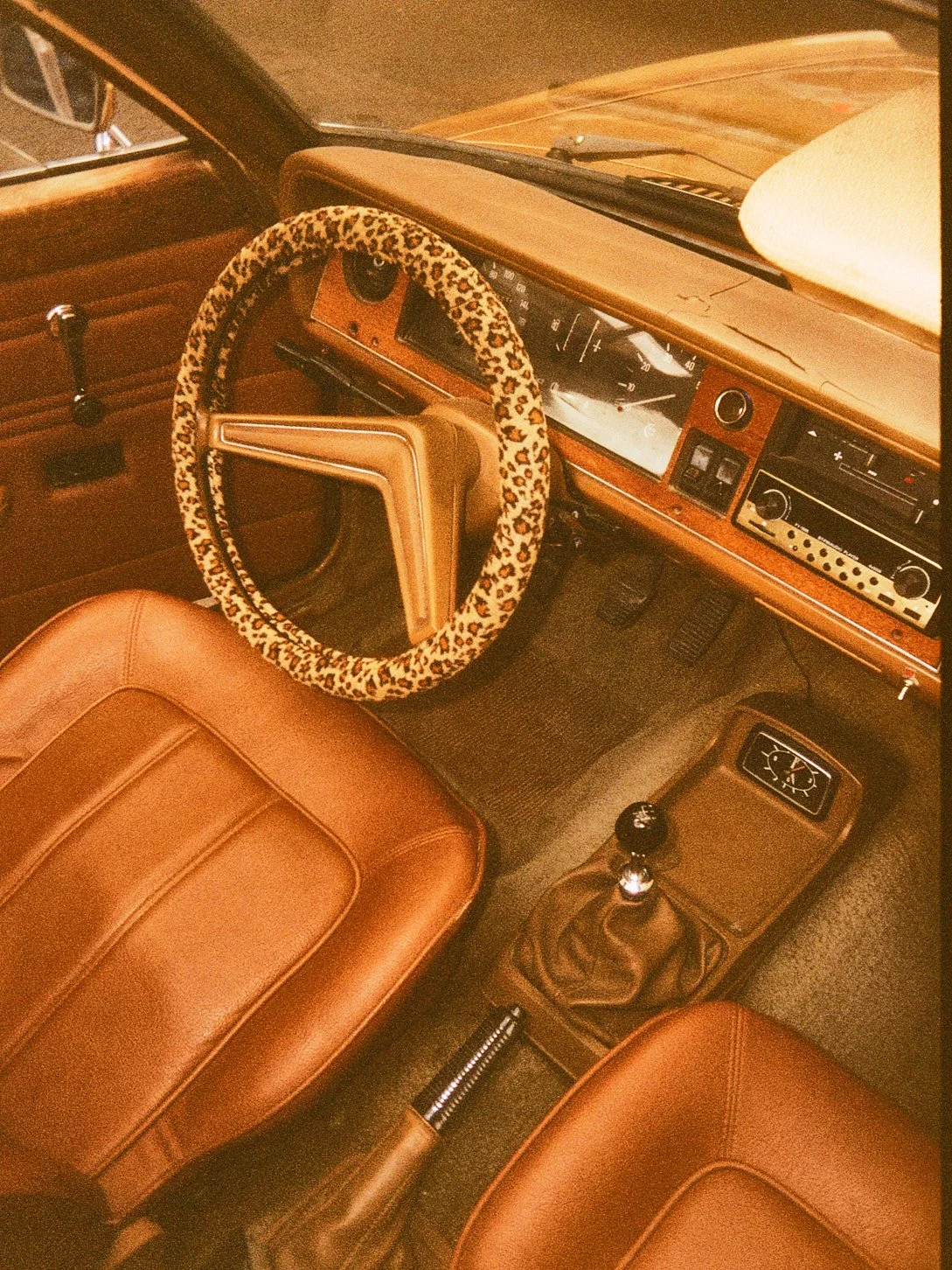 Ceci est l'intérieur d'une voiture vintage avec des sièges en cuir marron, un volant avec une housse à motif léopard, un tableau de bord en bois avec un système audio, un levier de vitesse, et un compteur horaire.