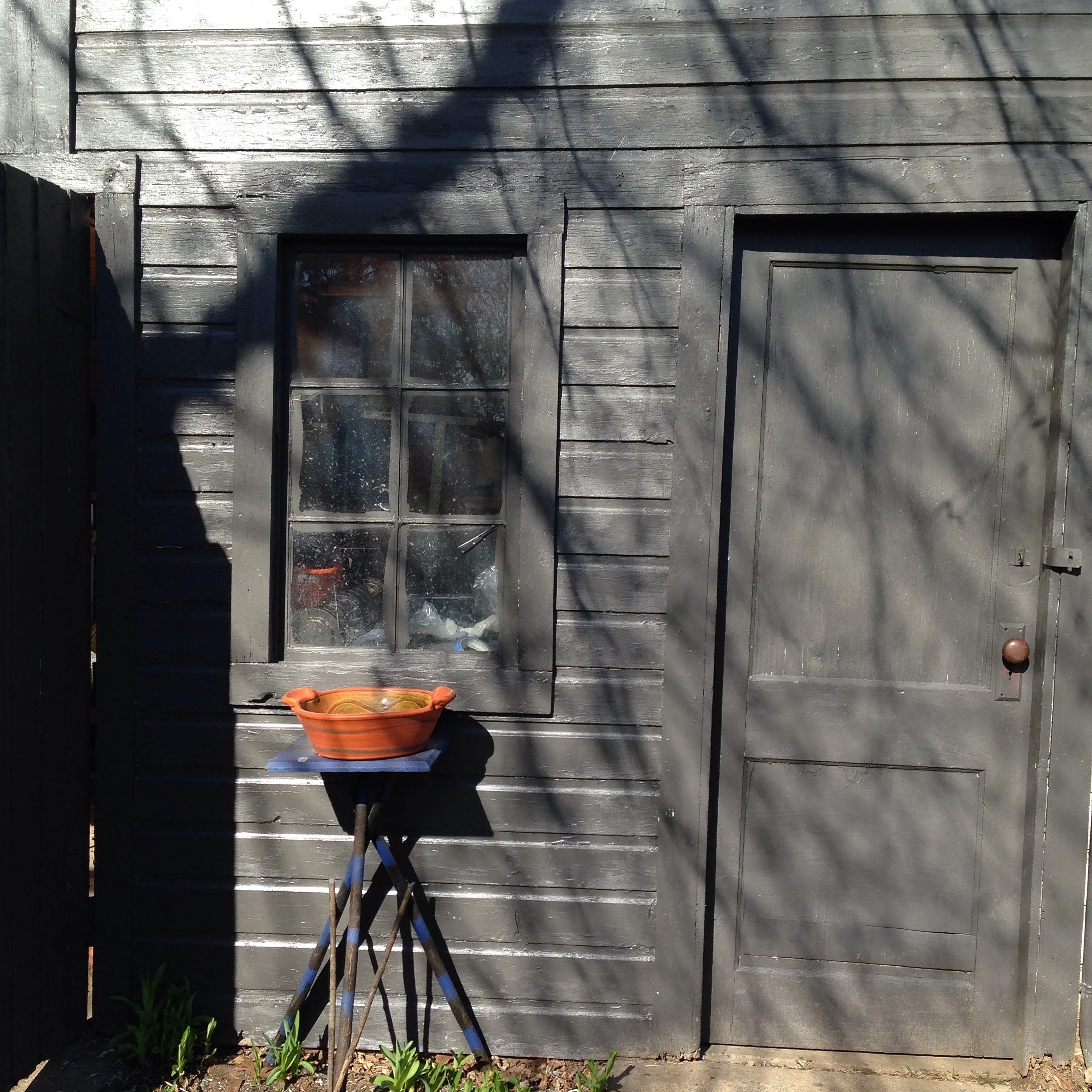 Exterior of a wooden building with gray painted walls, featuring a window and a door. A small orange planter sits on a stand in front of the window, with shadows of tree branches cast on the wall. Some green plants are visible at the bottom.