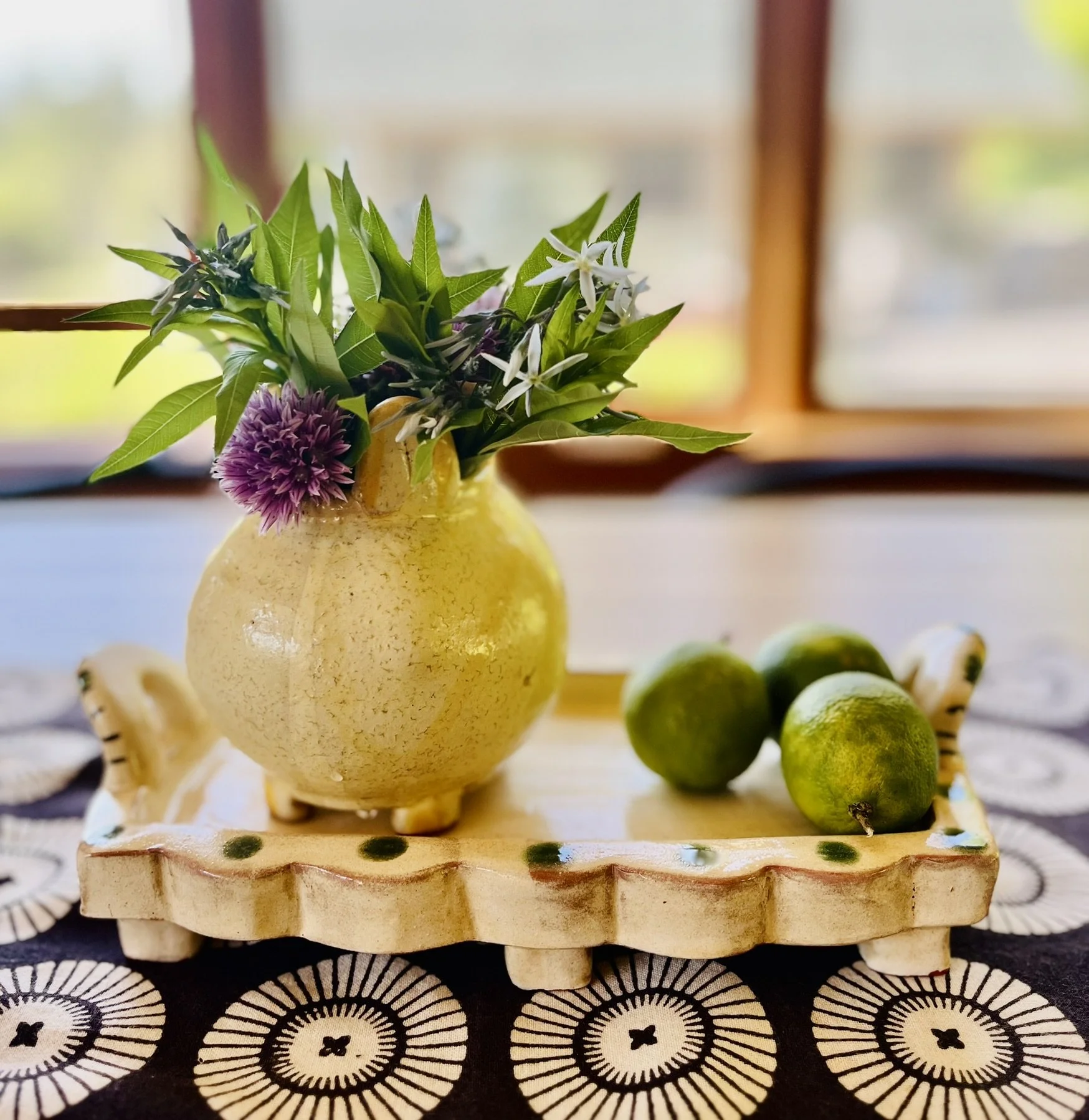 Decorative ceramic vase with purple and white flowers and green leaves on a tray with three limes, placed on a patterned tablecloth.