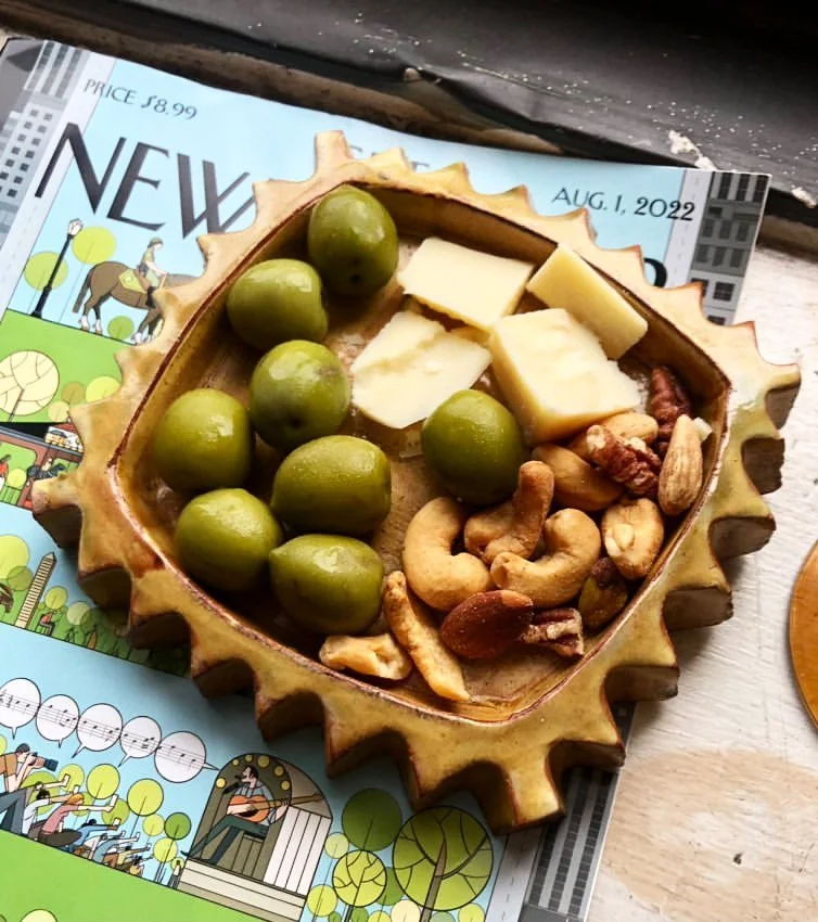 A decorative dish containing green olives, cheese slices, and assorted nuts on a table with a magazine in the background.