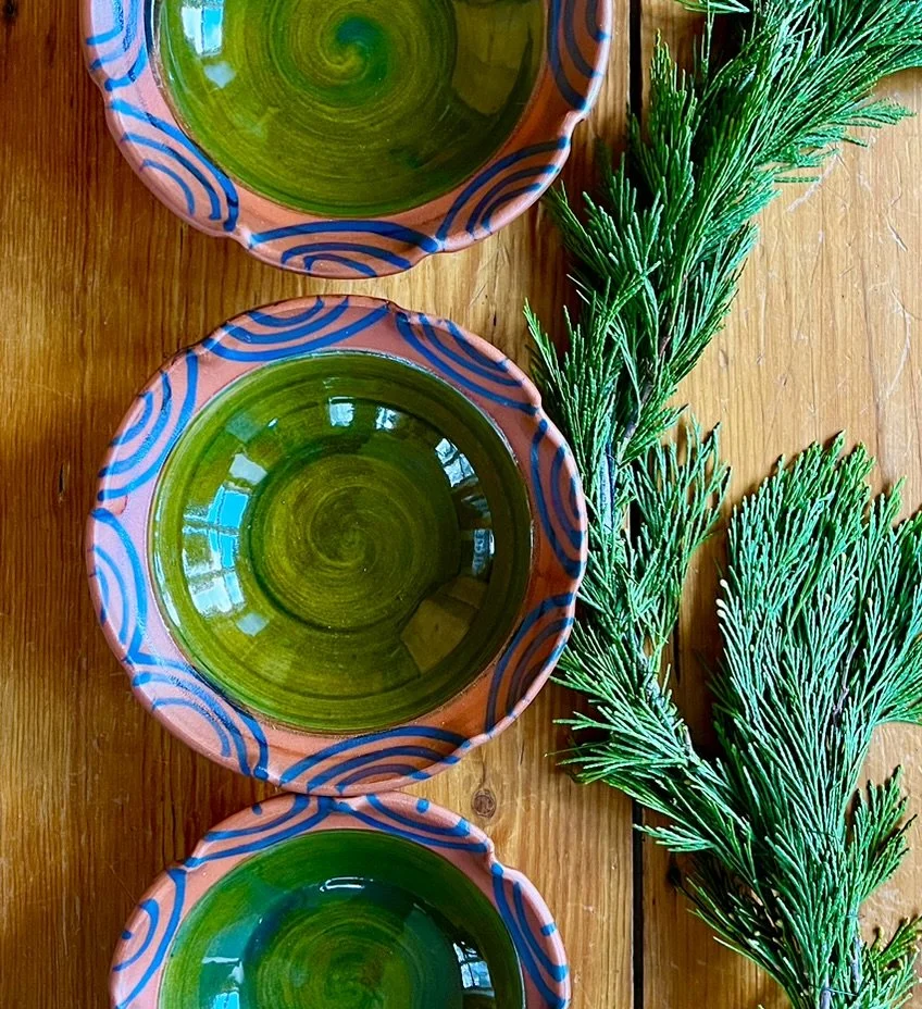 Three decorative ceramic bowls with green interiors and blue spiral designs, placed on a wooden surface next to green foliage.