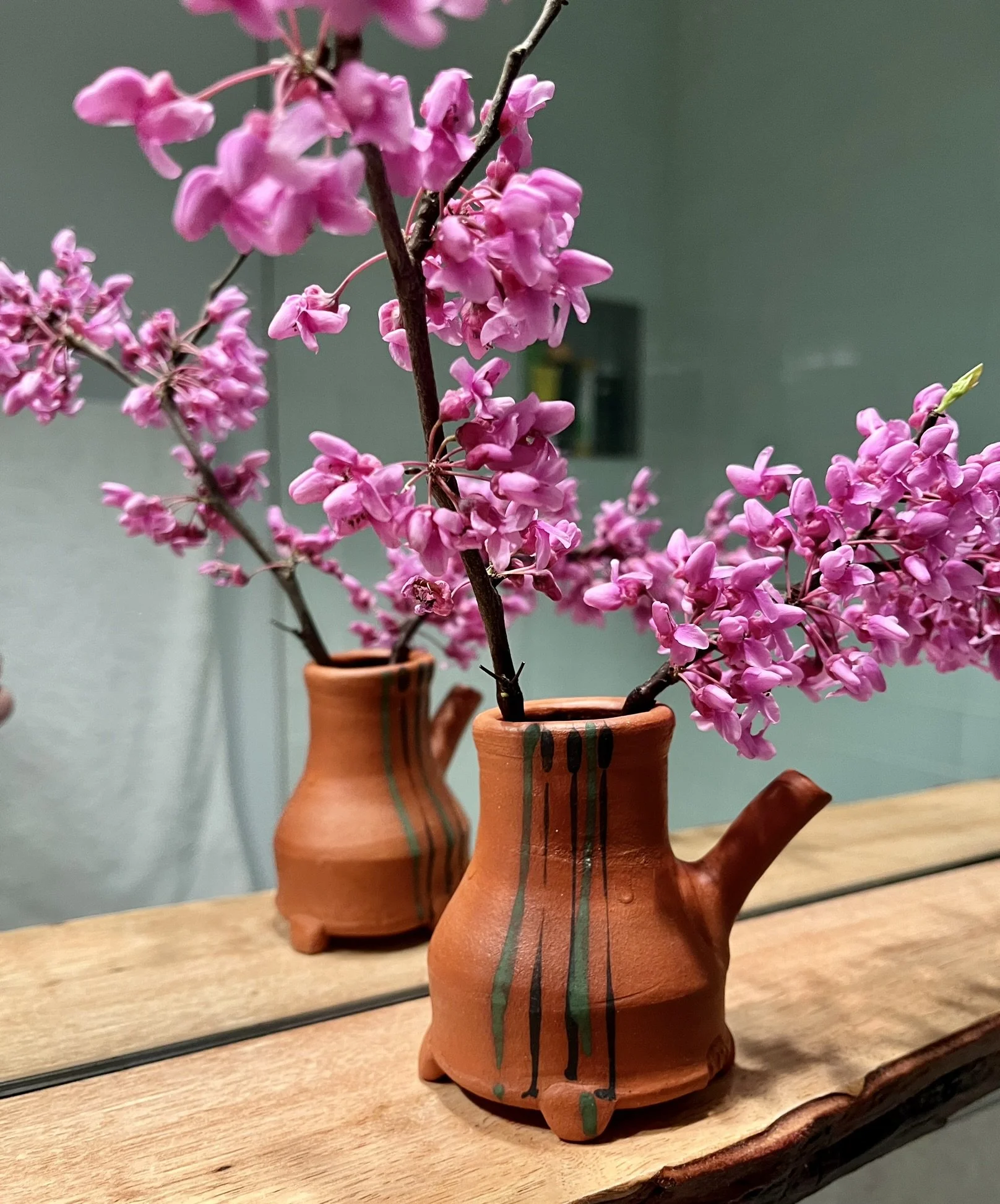 A terracotta vase with a side spout, containing branches of pink flowers, reflected in a mirror on a wooden surface.