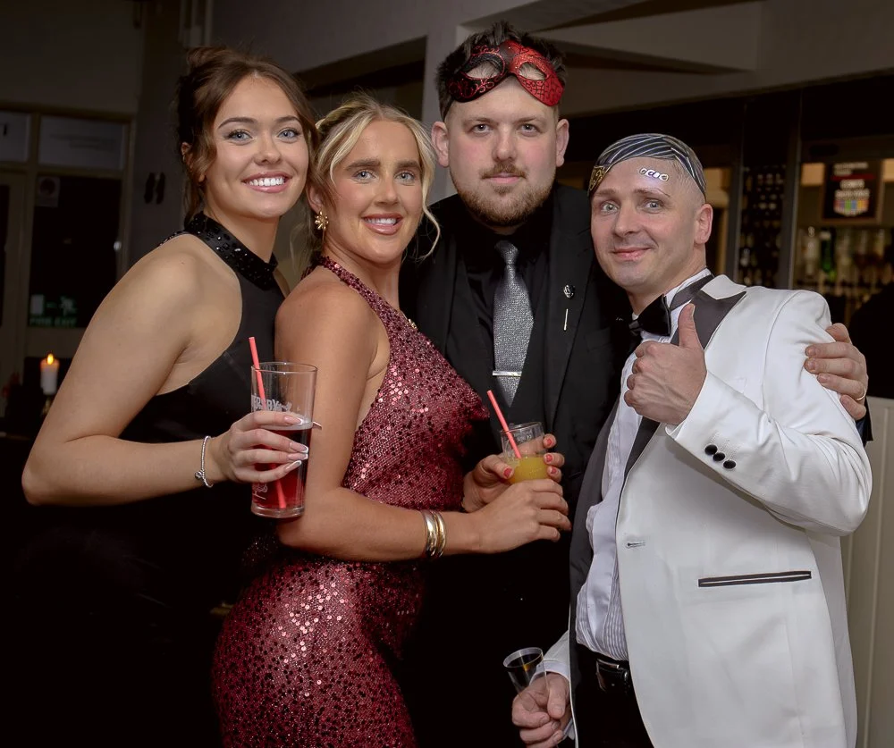 Four people dressed in formal attire celebrating at a party, holding drinks, with one person giving a thumbs-up.